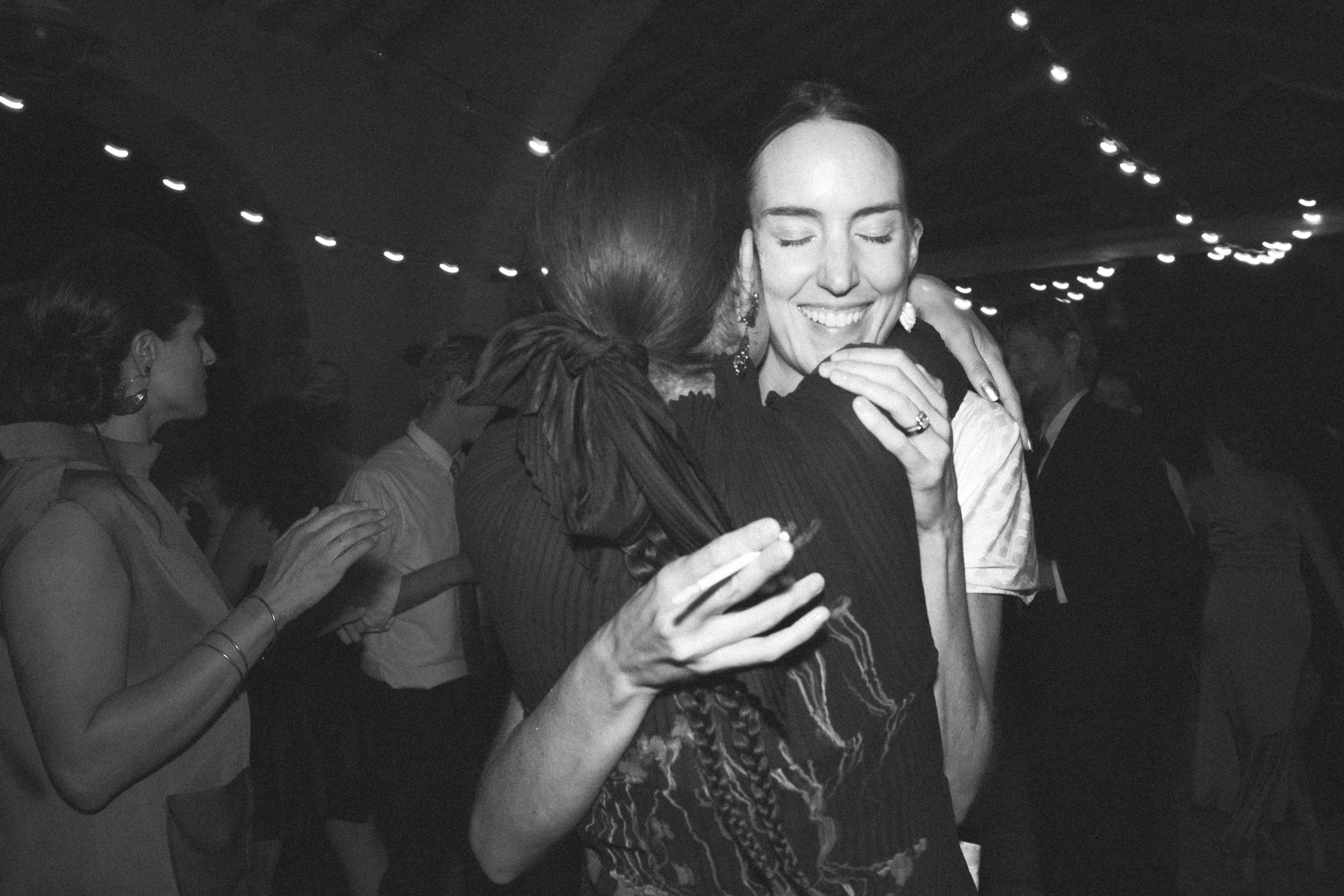 Two women hugging, smiling with eyes closed, in a social gathering or dance event, with other people and string lights in the background.