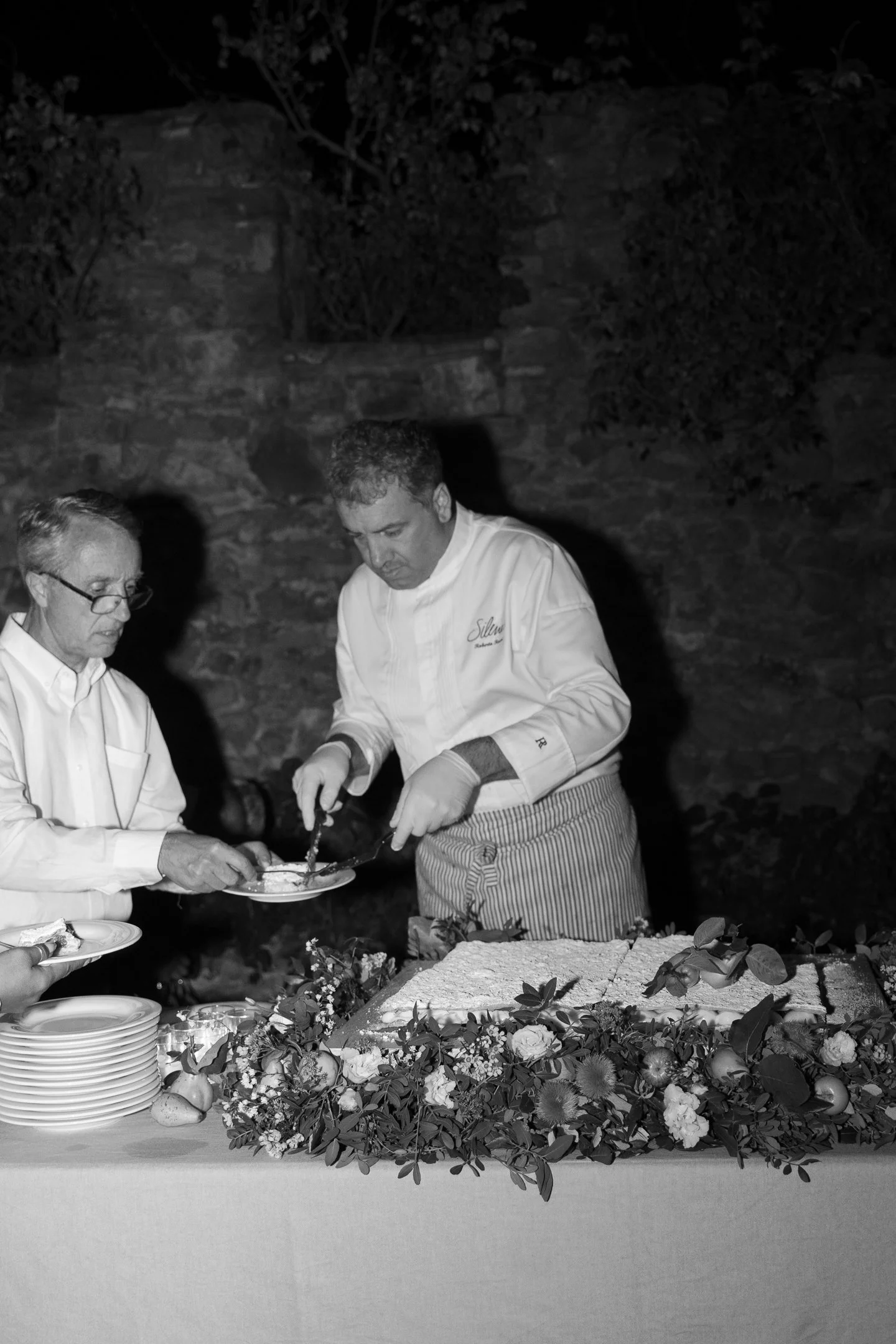 Two chefs serving dessert at a table decorated with flowers and fruit, in an outdoor setting at night.