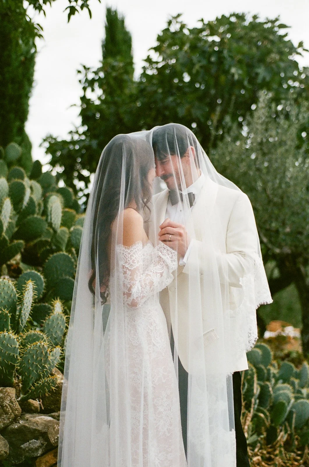 A bride and groom are standing close together outdoors, under a veil, with cacti and trees in the background, sharing an intimate moment.