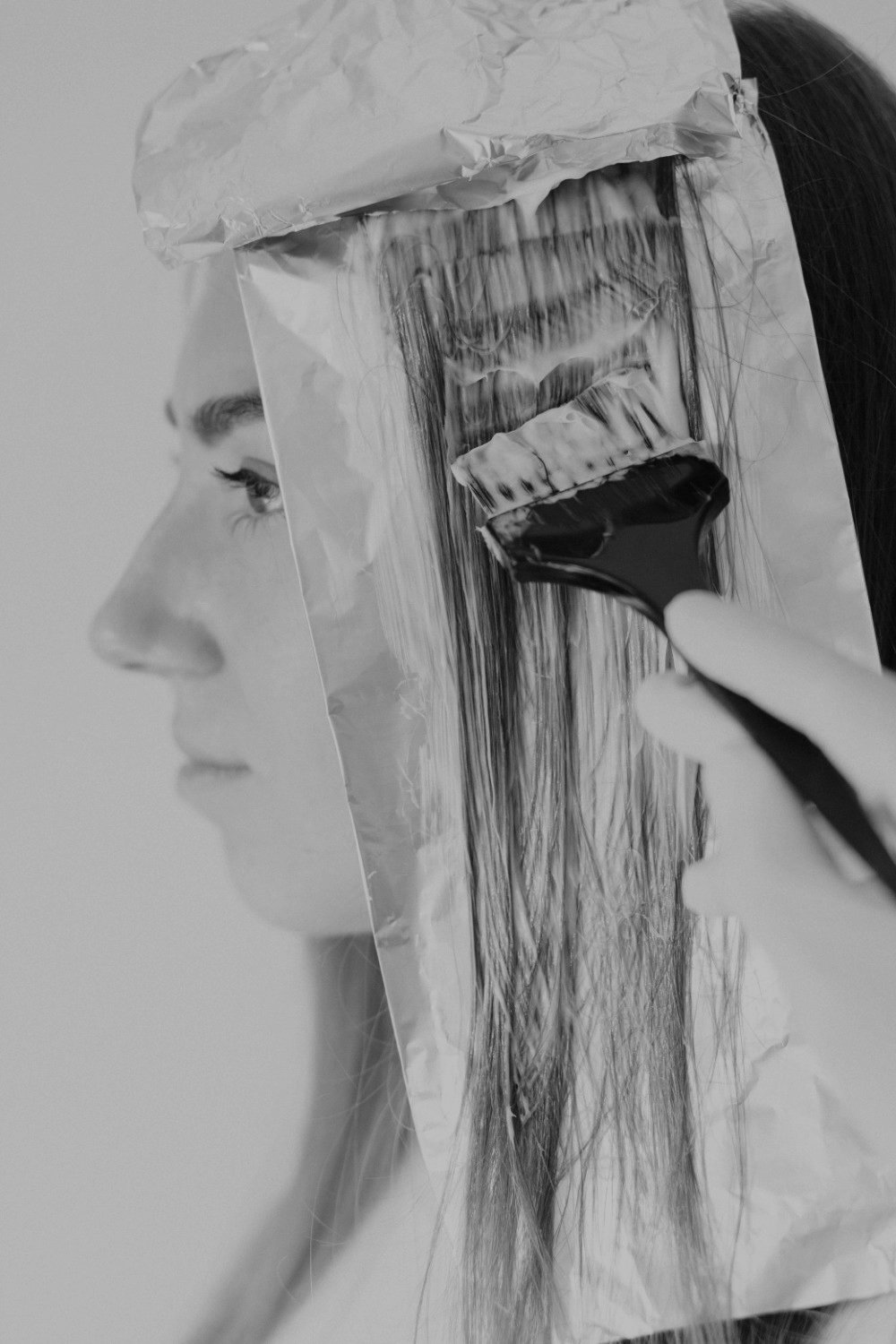 A woman with makeup is holding a spatula and applying black hair dye to her long hair, which is covered with a plastic cape for protection.