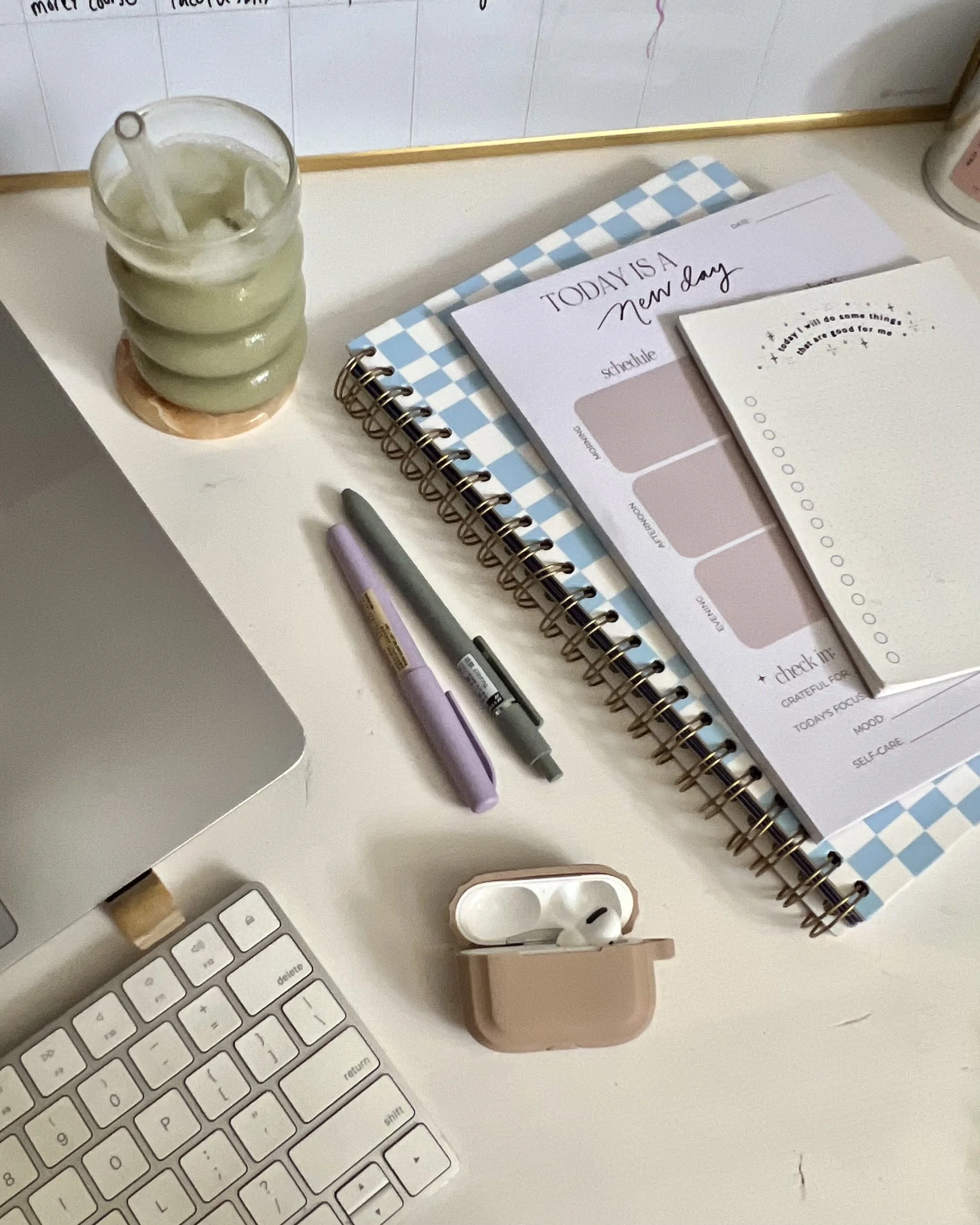 An organized workspace with a white keyboard, a pen, a small pink case, and sticky notes on a white desk. There is a glass of water with ice and a straw, a planner or journal, and additional stationery items on the desk.