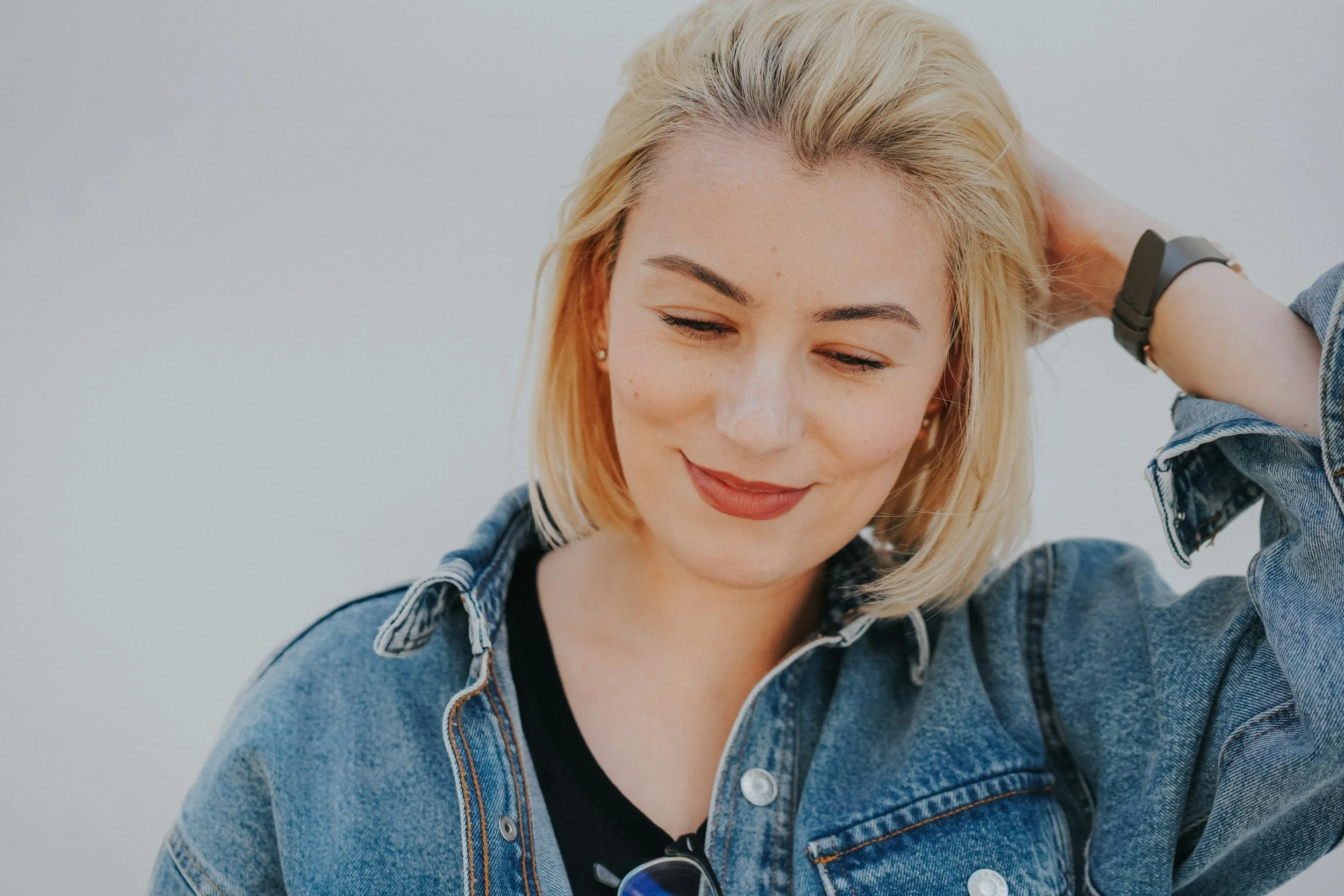 Close-up photo of a smiling woman with blonde hair, wearing a denim jacket, looking down, with her right hand raised and resting on her head.
