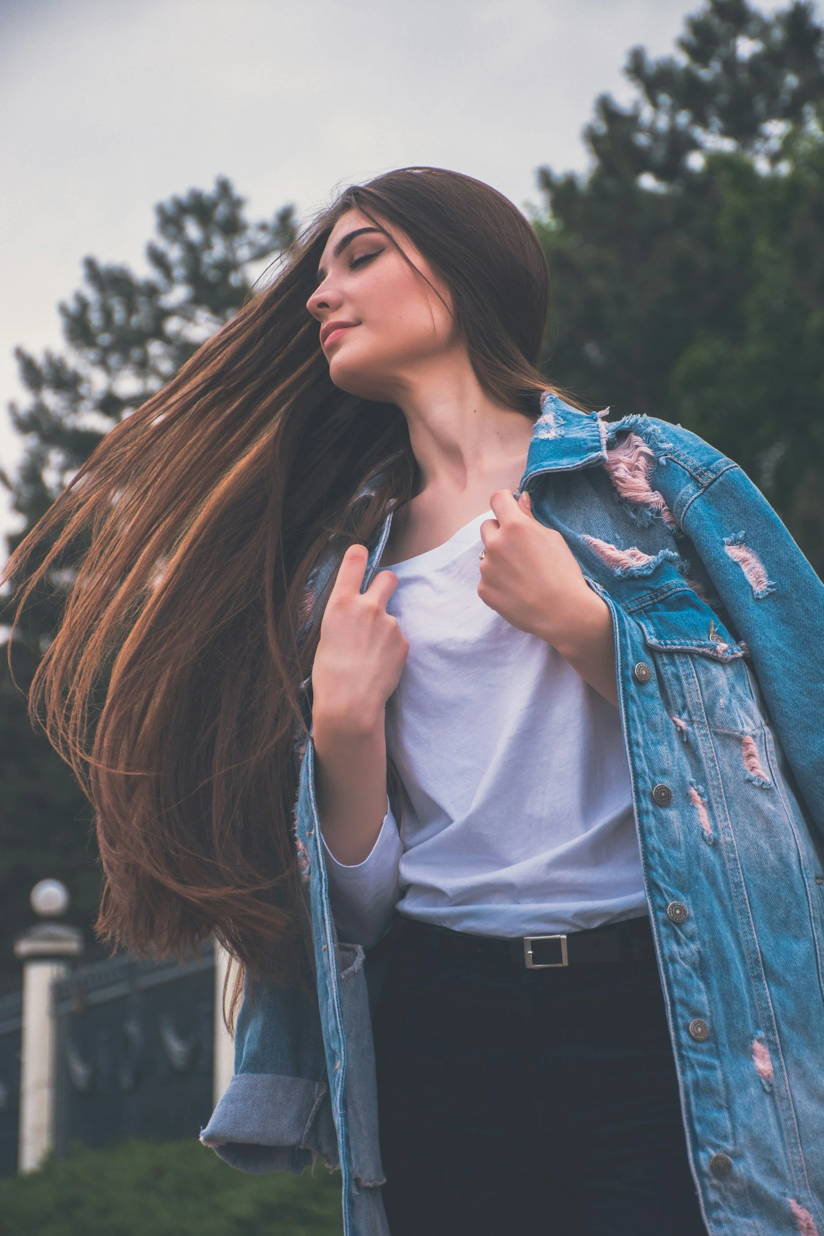 Young woman with long brown hair, wearing a white shirt and a distressed denim jacket, standing outdoors with trees in the background, eyes closed, and her hand holding the collar of her jacket.