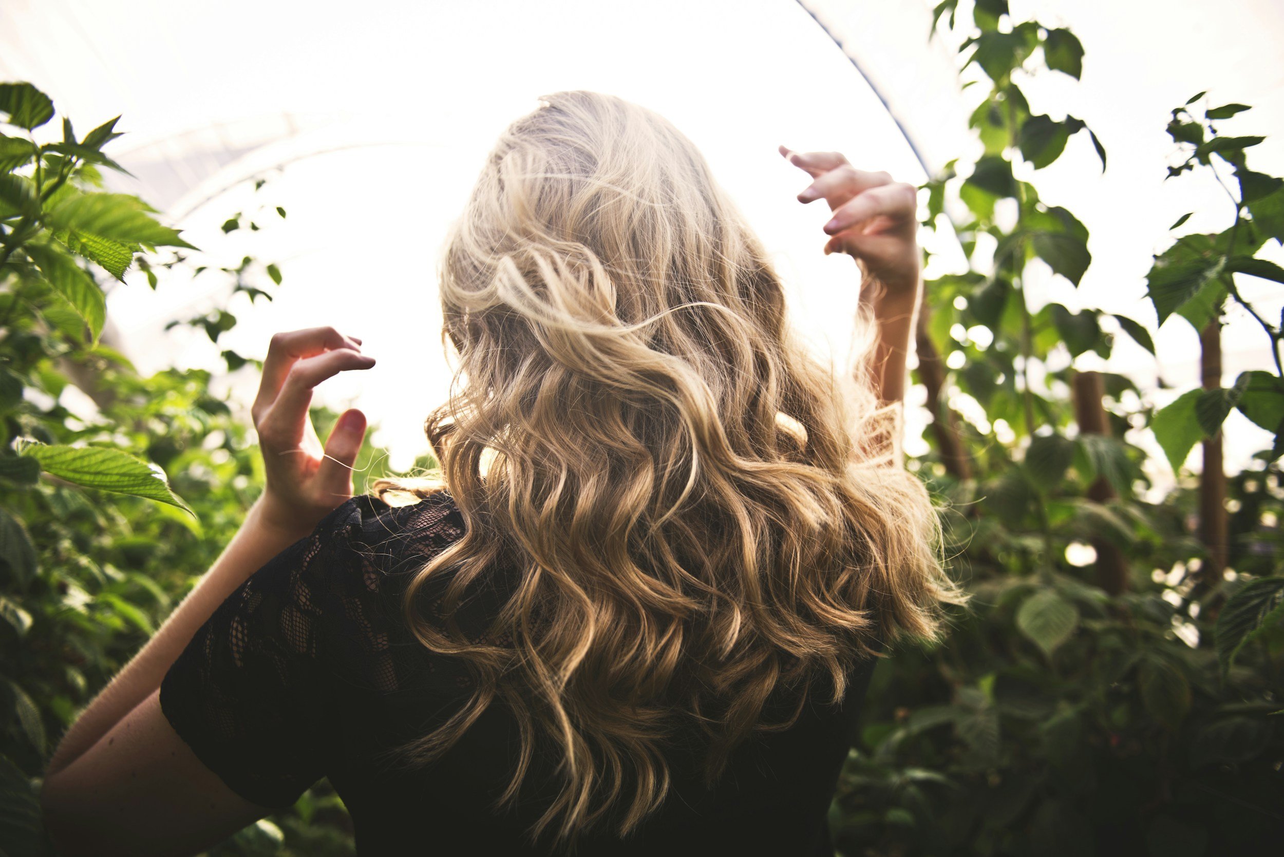 A woman with long, curly blonde hair standing among green plants or trees, holding an umbrella with her left hand.