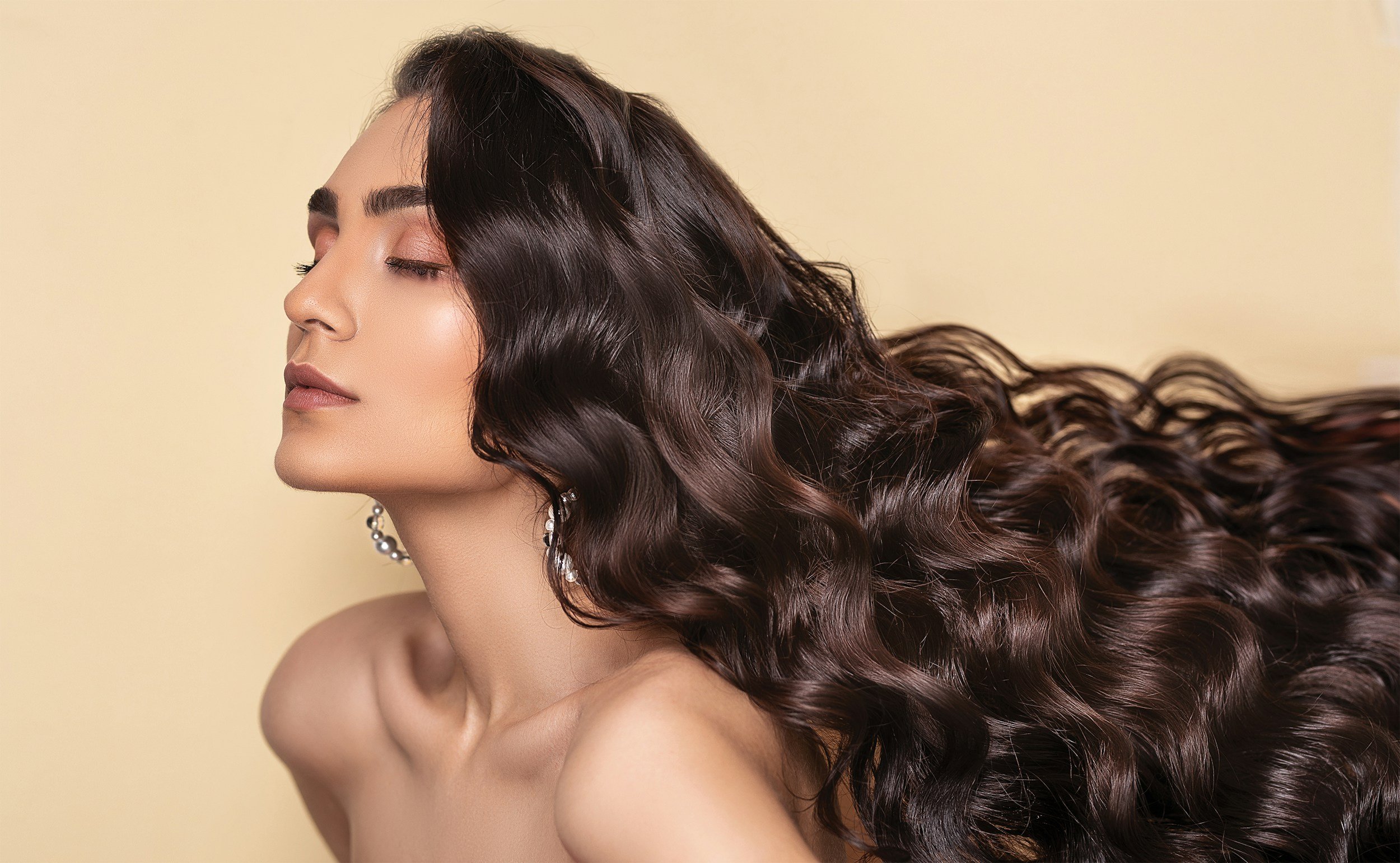 A woman with long, wavy dark brown hair and closed eyes, wearing pearl earrings, against a plain yellow background.