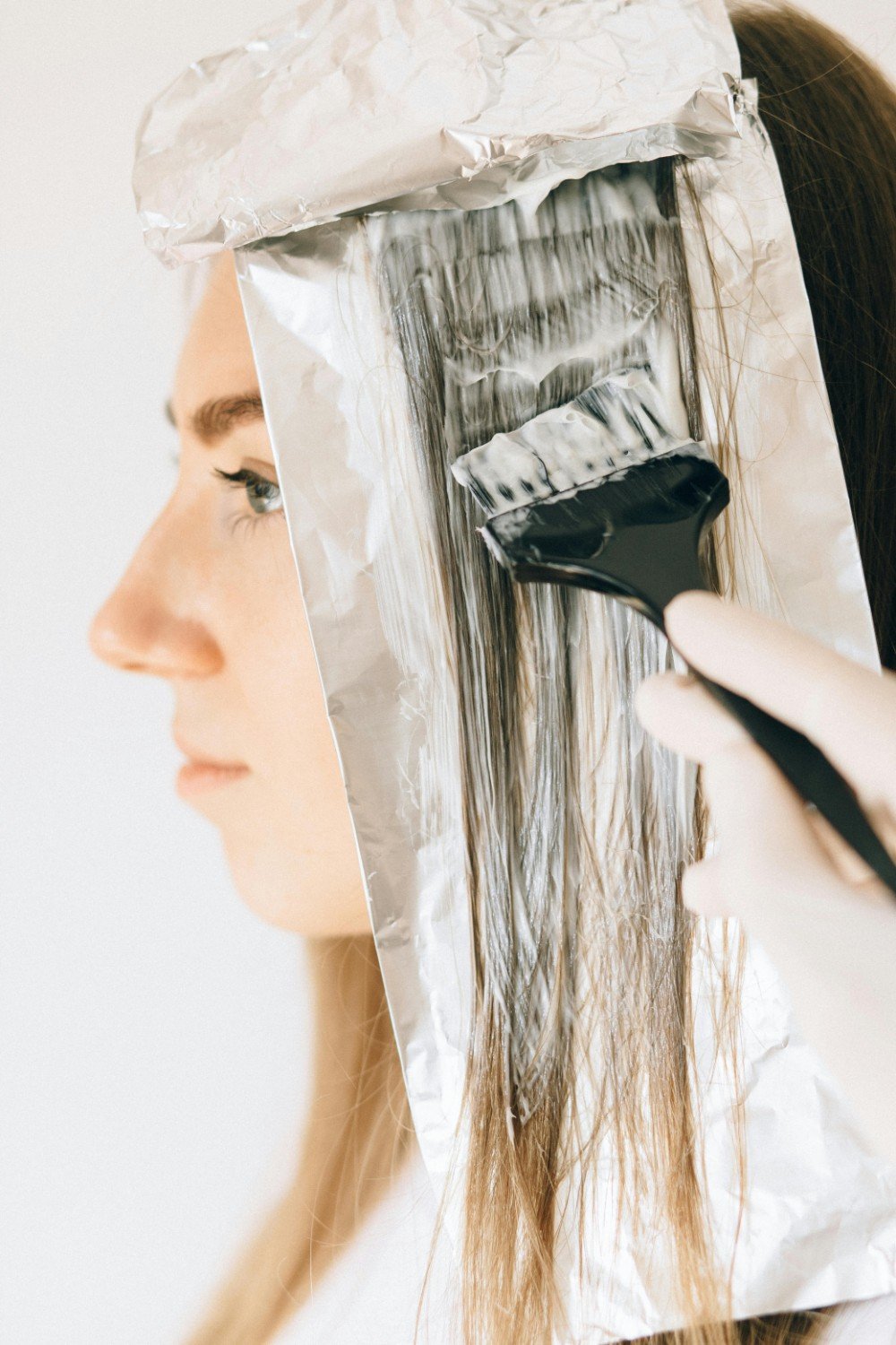 A woman is dyeing her hair using black hair dye applied with a brush onto a foil sheet. Her face is in profile, and she has long brown hair and makeup with eyeliner.
