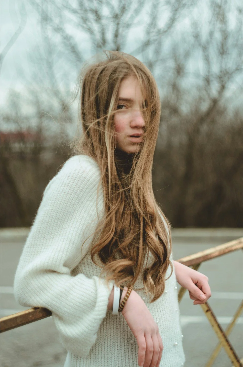 A young woman with long, light brown hair, wearing a white knitted sweater and black turtleneck, standing outdoors near a rusty railing with leafless trees in the background.