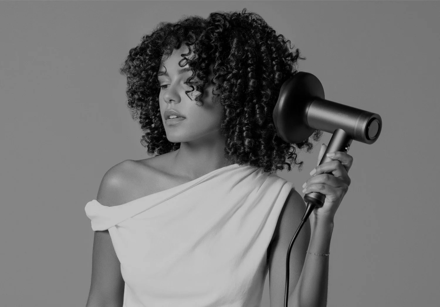 A woman with curly hair holding a hair dryer against her head, wearing an off-shoulder white top, with a neutral expression on her face.