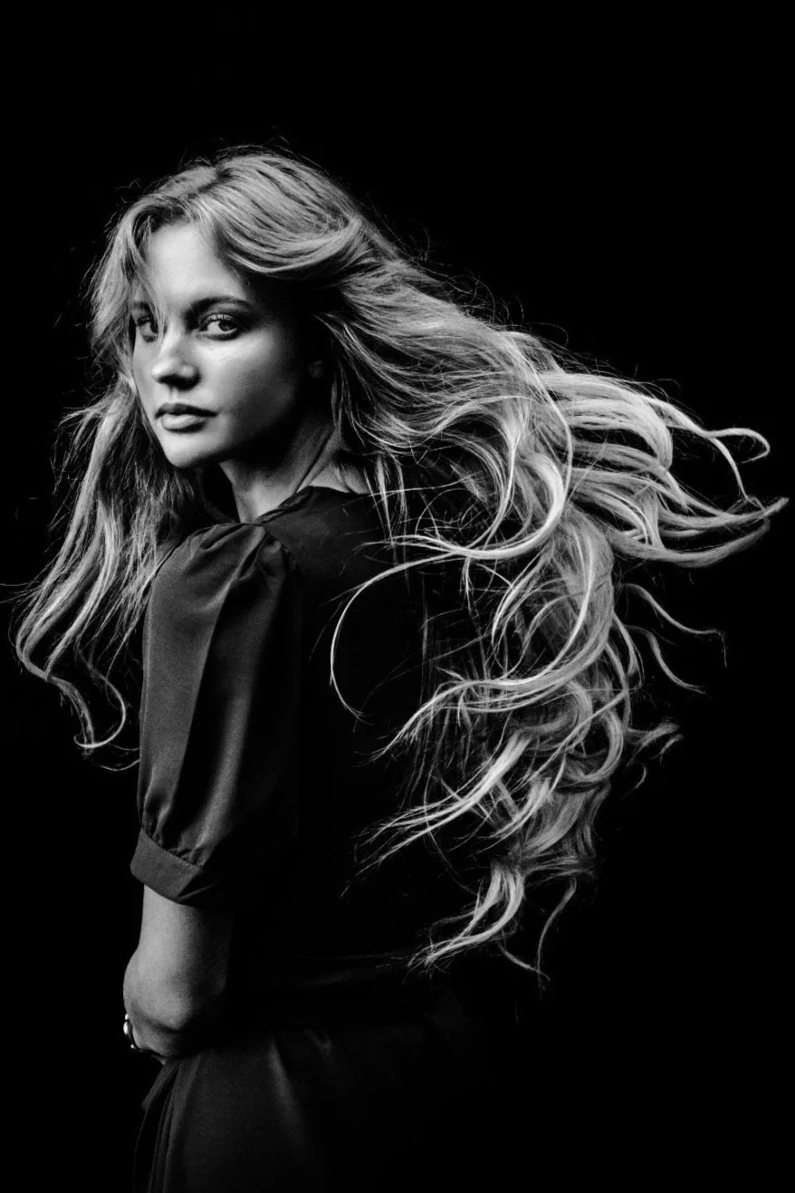 Black and white portrait of a woman with long, wavy hair, looking over her shoulder, wearing a dark blouse with puffed sleeves, against a dark background.