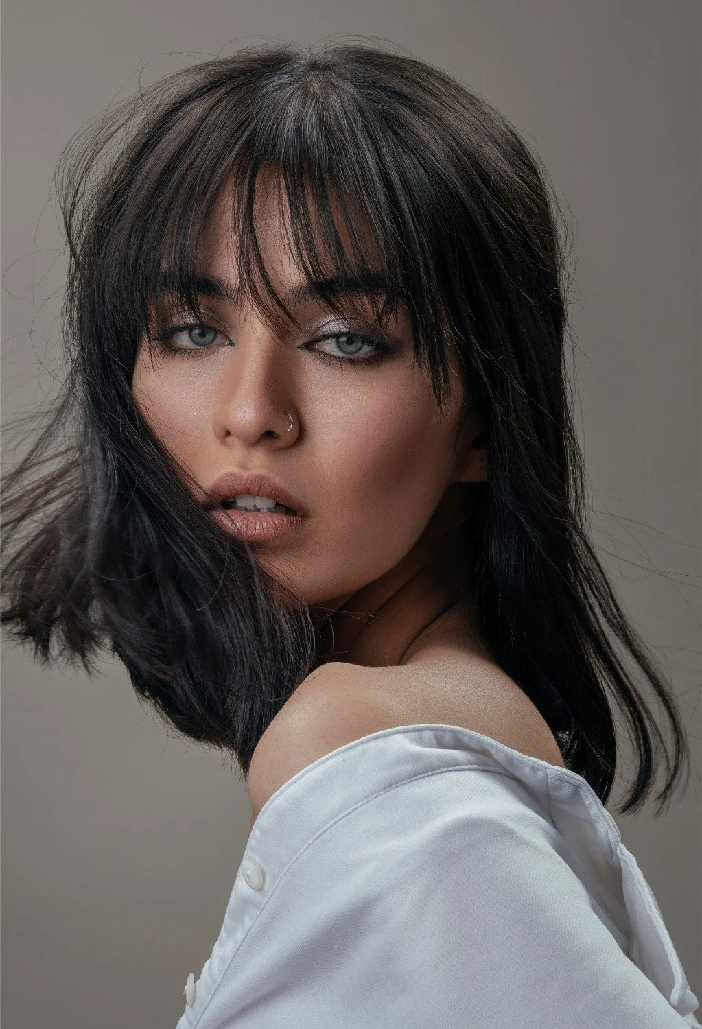 A young woman with dark, messy hair, light skin, blue eyes, and a nose ring, wearing a white shirt, looking over her shoulder with a neutral expression.
