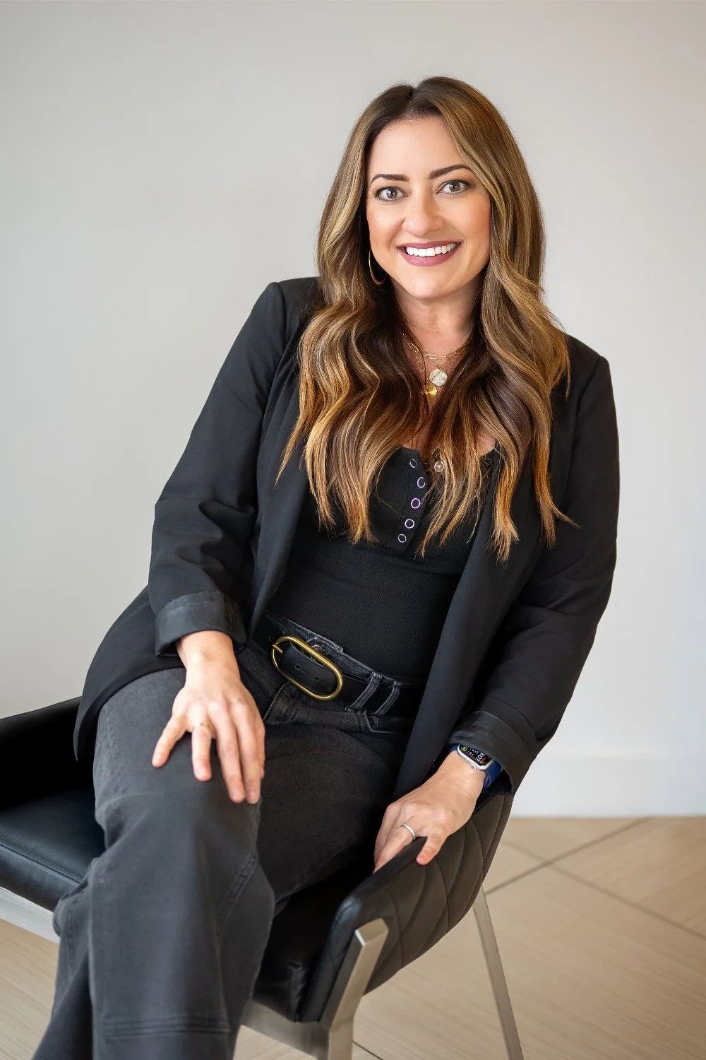 A woman with long, wavy brown hair and a bright smile, wearing a black blazer and black pants, sitting on a black chair against a plain white background.