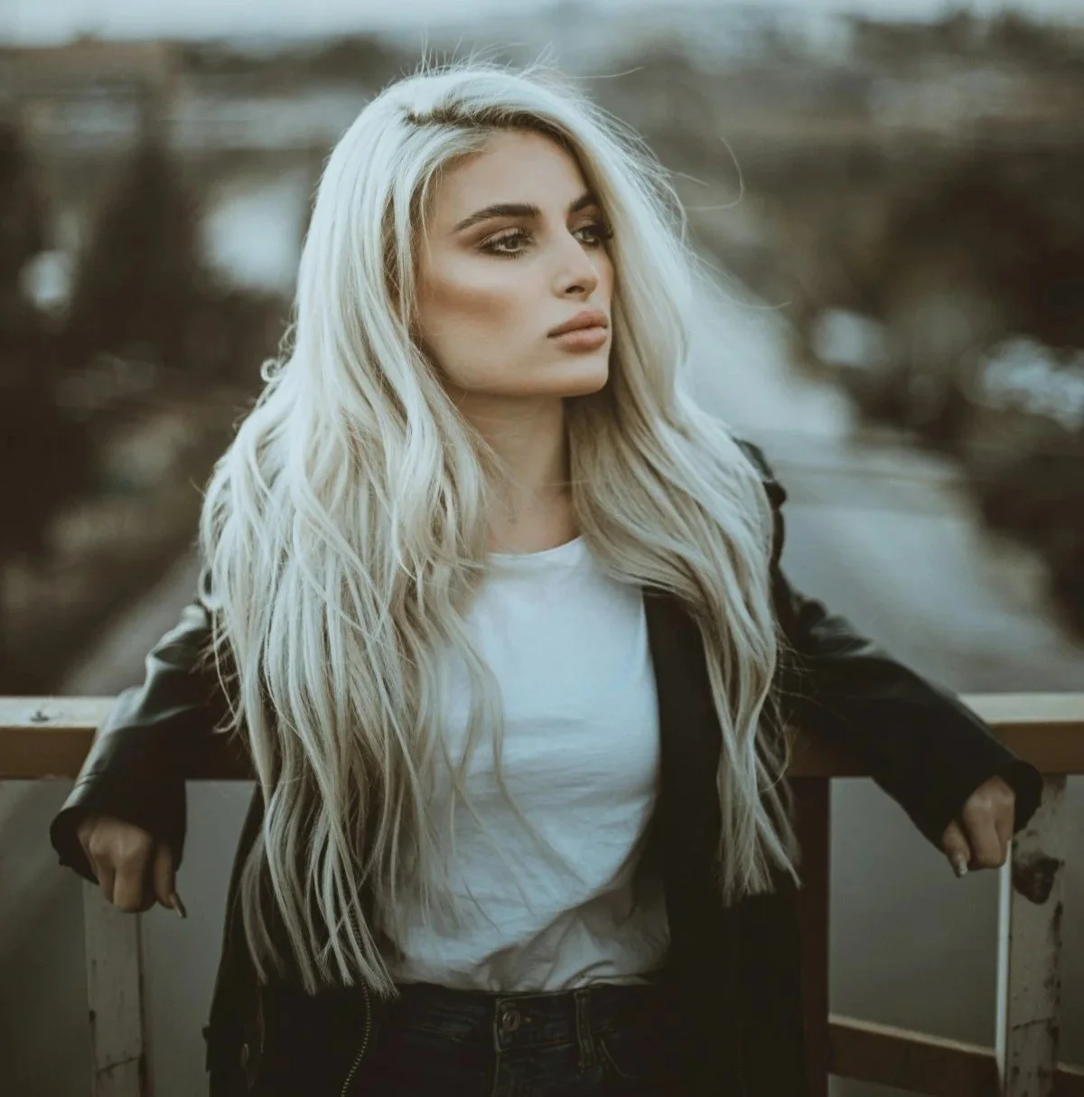 A woman with long wavy platinum blonde hair, wearing a white t-shirt and black jacket, sitting outdoors on a wooden railing, looking to the right with a serious expression.