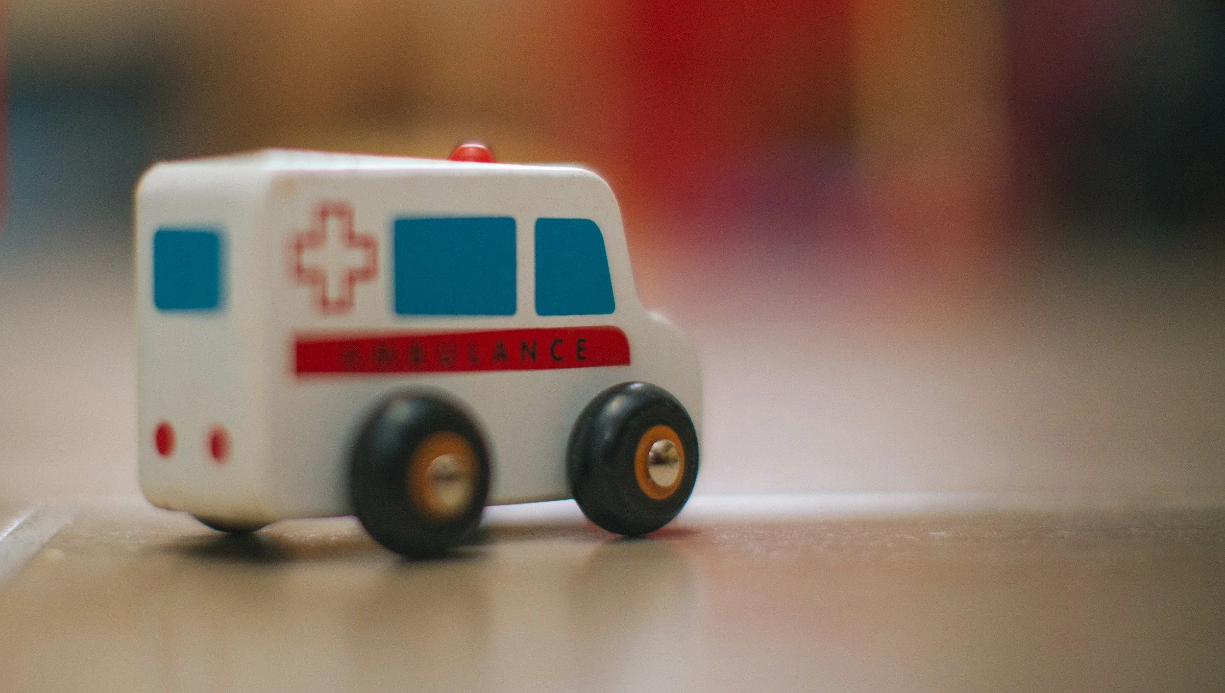A small toy ambulance sitting on a wooden surface with a blurred colorful background.