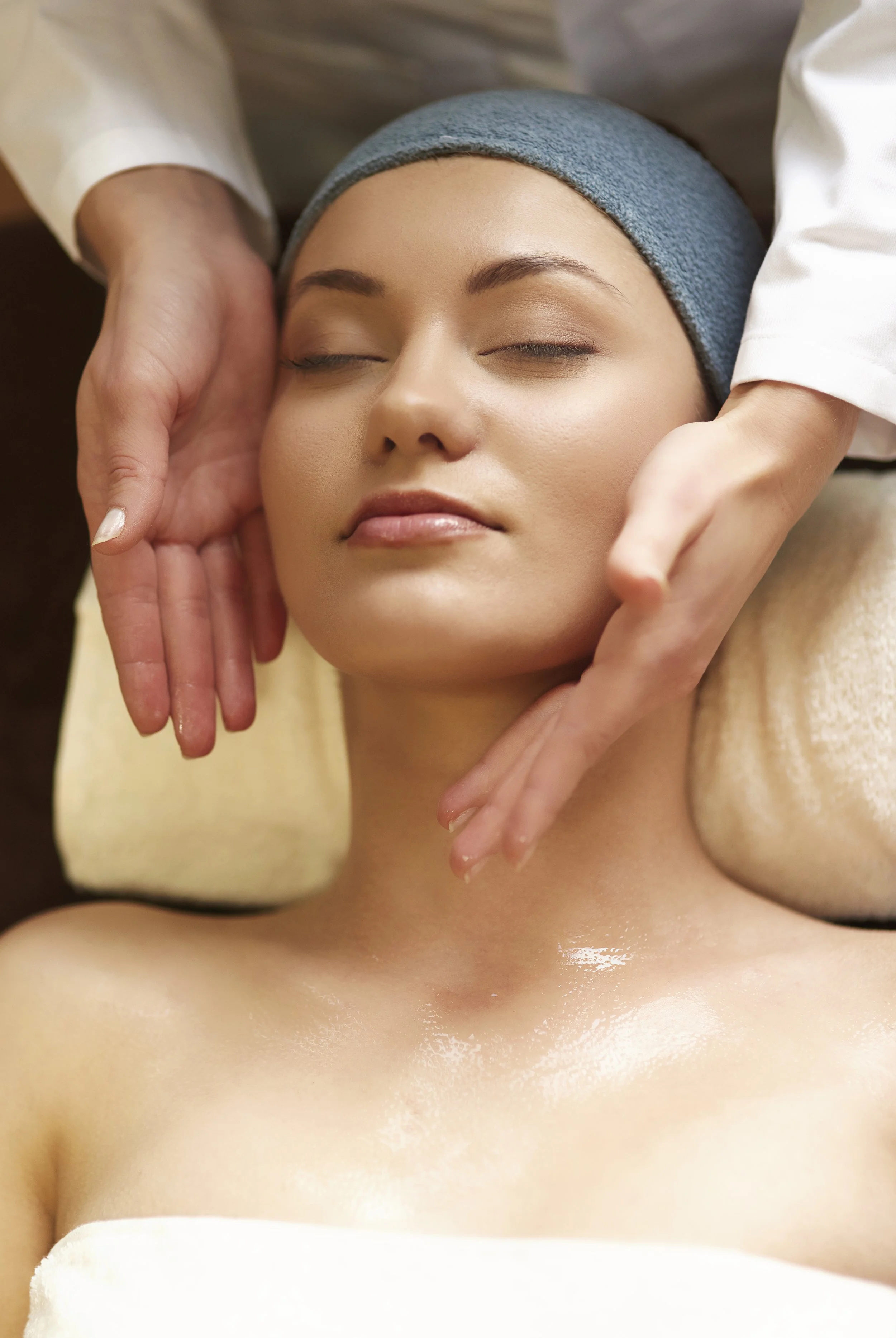 A woman receiving a face massage or facial treatment, lying down with eyes closed, wearing a blue headband.
