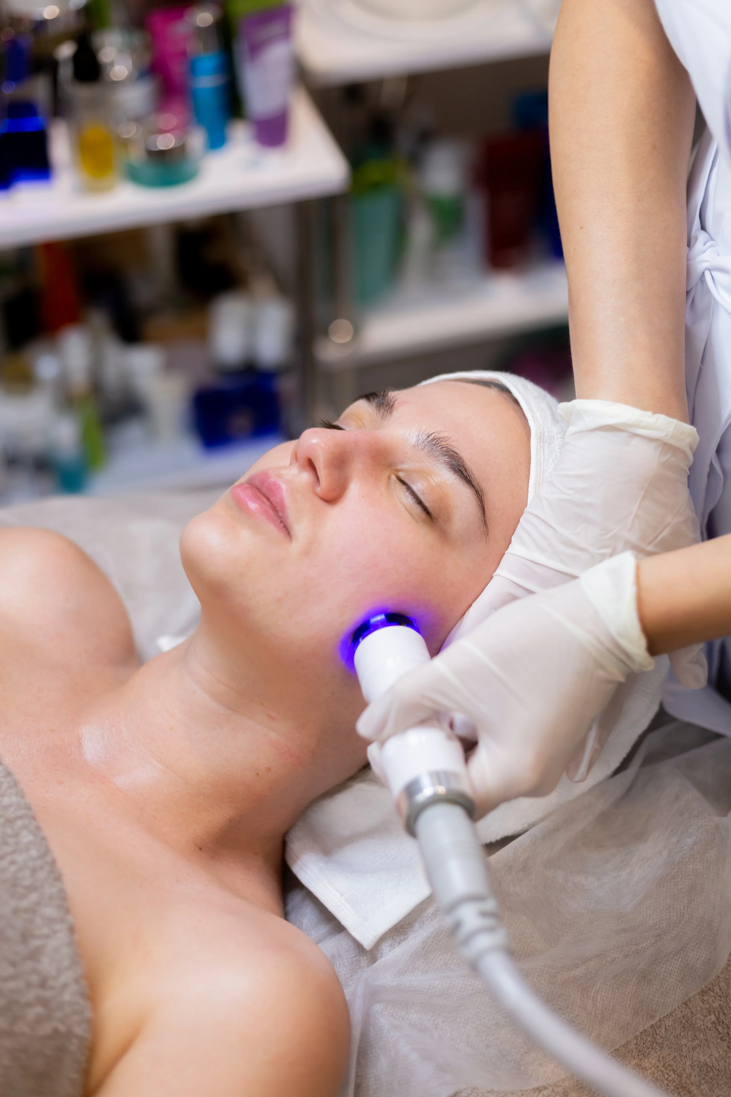 Person receiving a facial treatment in a spa or clinic setting, lying on their back with eyes closed while a professional uses technology on their face.