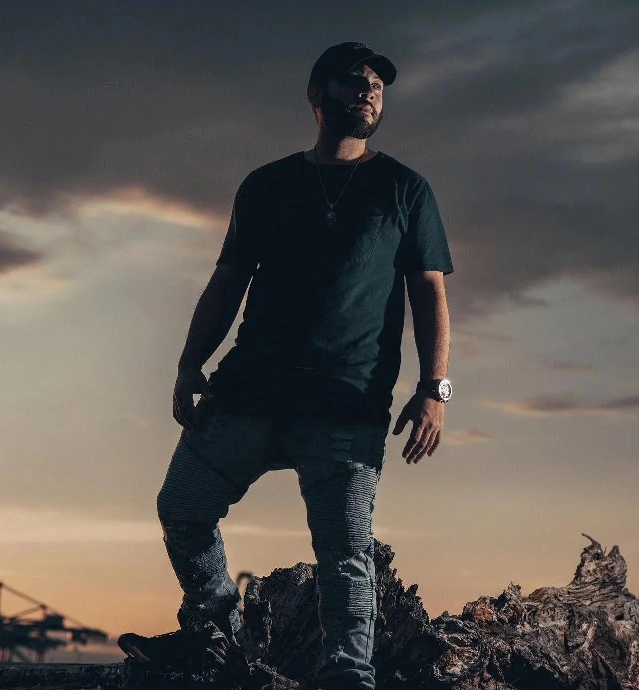 A man standing outdoors on a piece of dead wood at sunset, dressed in black clothing, wearing a watch and necklace, with a cloudy sky in the background.