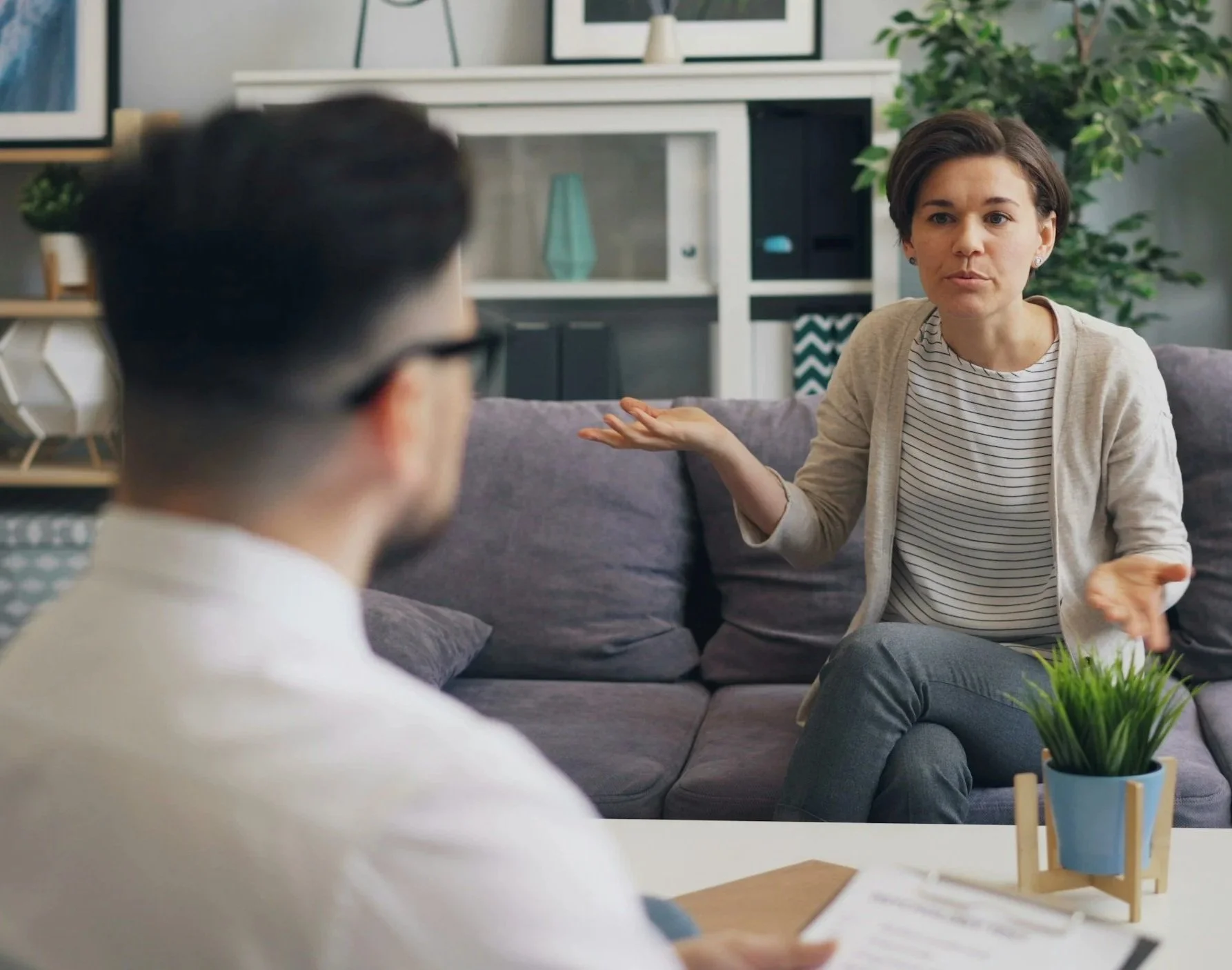 Woman in a beige cardigan and striped shirt gesturing while talking to man in glasses in a living room with plants and framed art.