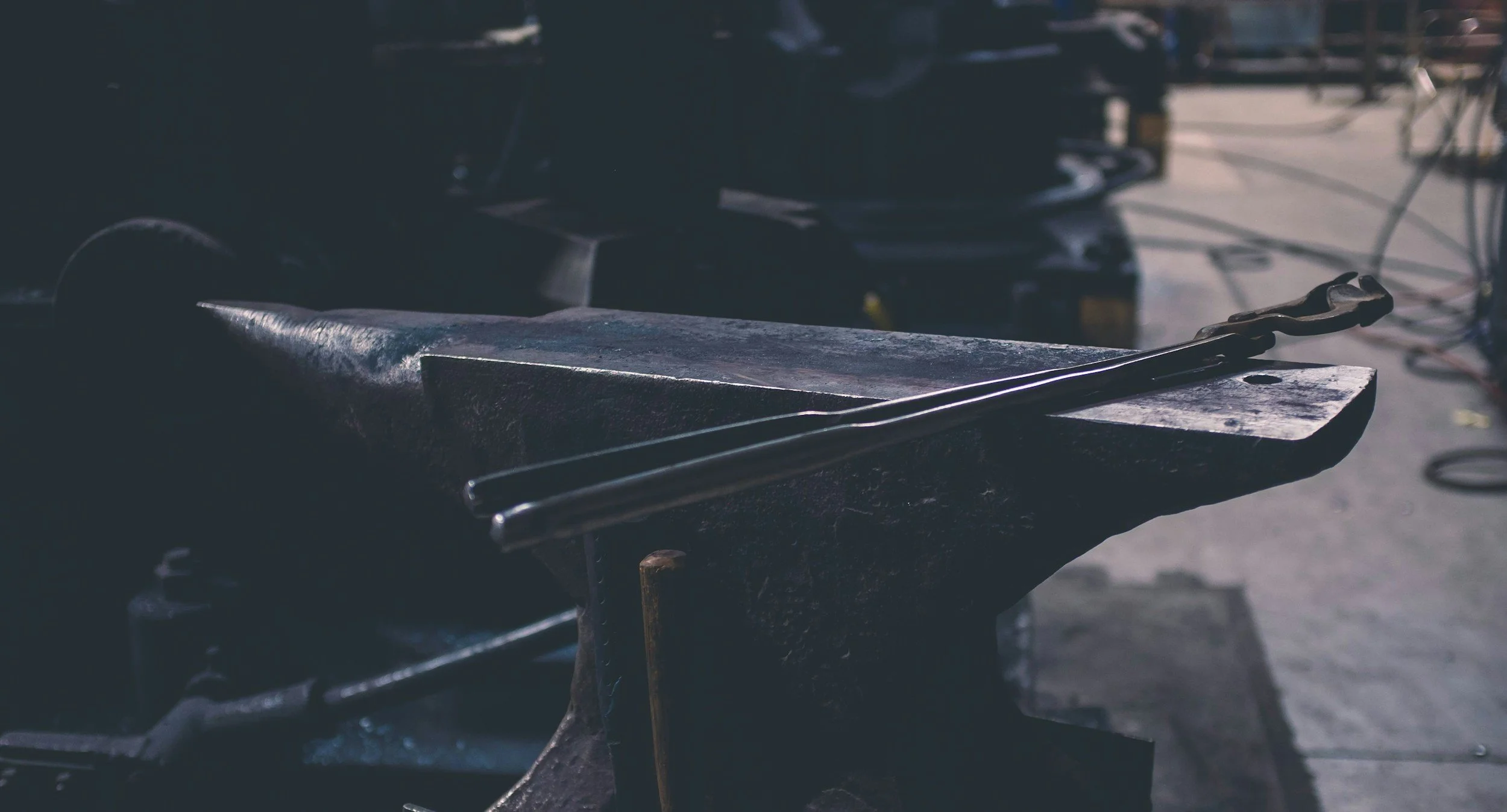 A close-up of a blacksmith's anvil with blacksmithing tongs resting on top, set in a workshop environment.