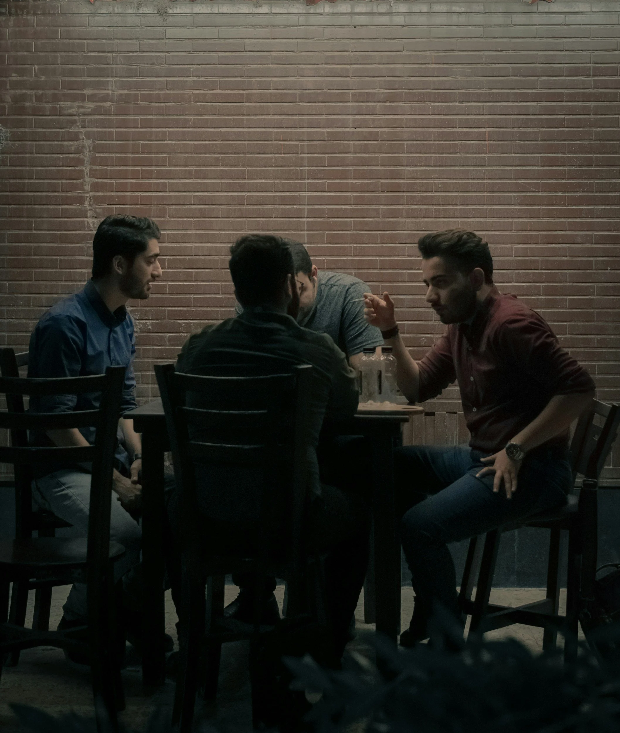 Four young men sitting around a table and engaging in conversation in a dimly lit room with a exposed brick wall in the background.