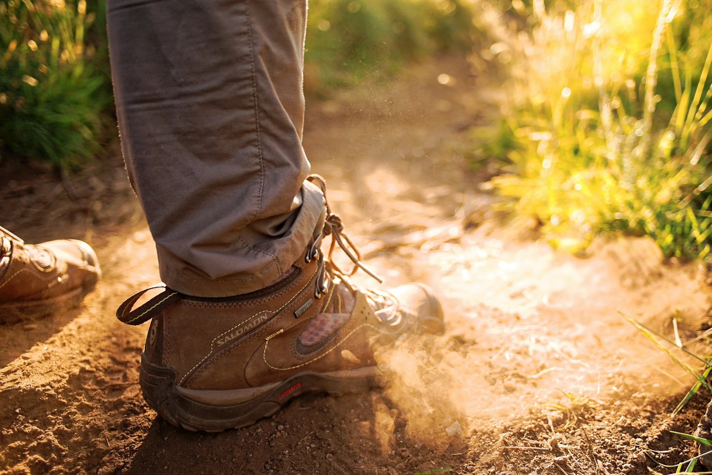 Close-up of a person's hiking boot on a dirt trail, with a pant leg visible and sunlight glowing from behind tall grasses.