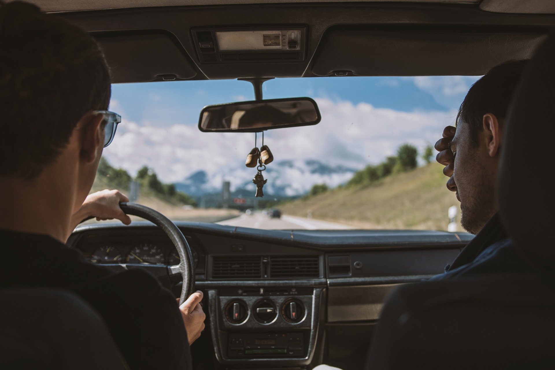 Photo taken from inside a car showing two passengers, one driving and one sitting in the front passenger seat, with a view of a sunny highway with green hills and trees outside.