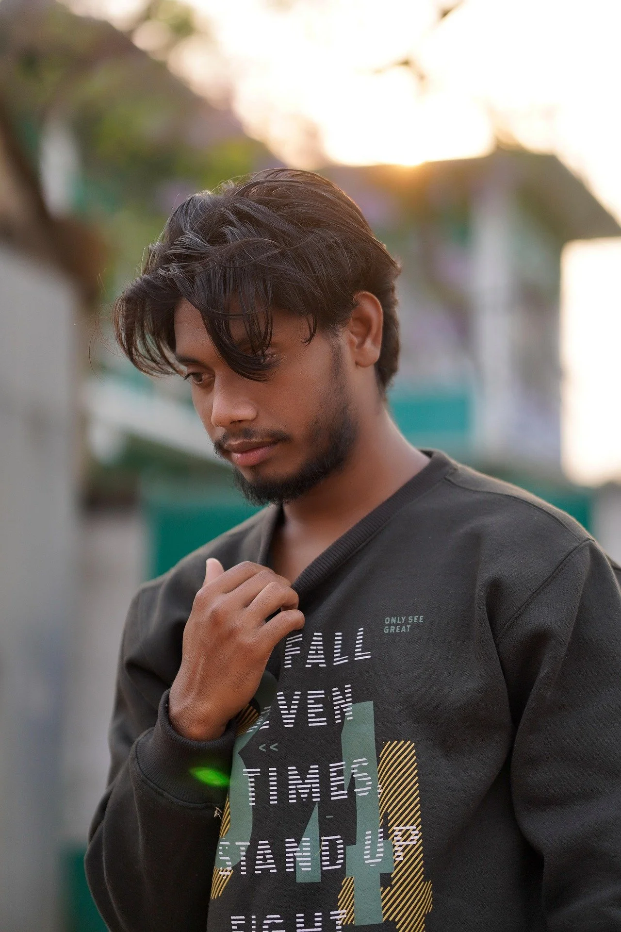 A young man with dark hair and beard looking down while holding his shirt collar with his right hand outdoors during sunset with blurred greenery and a building in the background.
