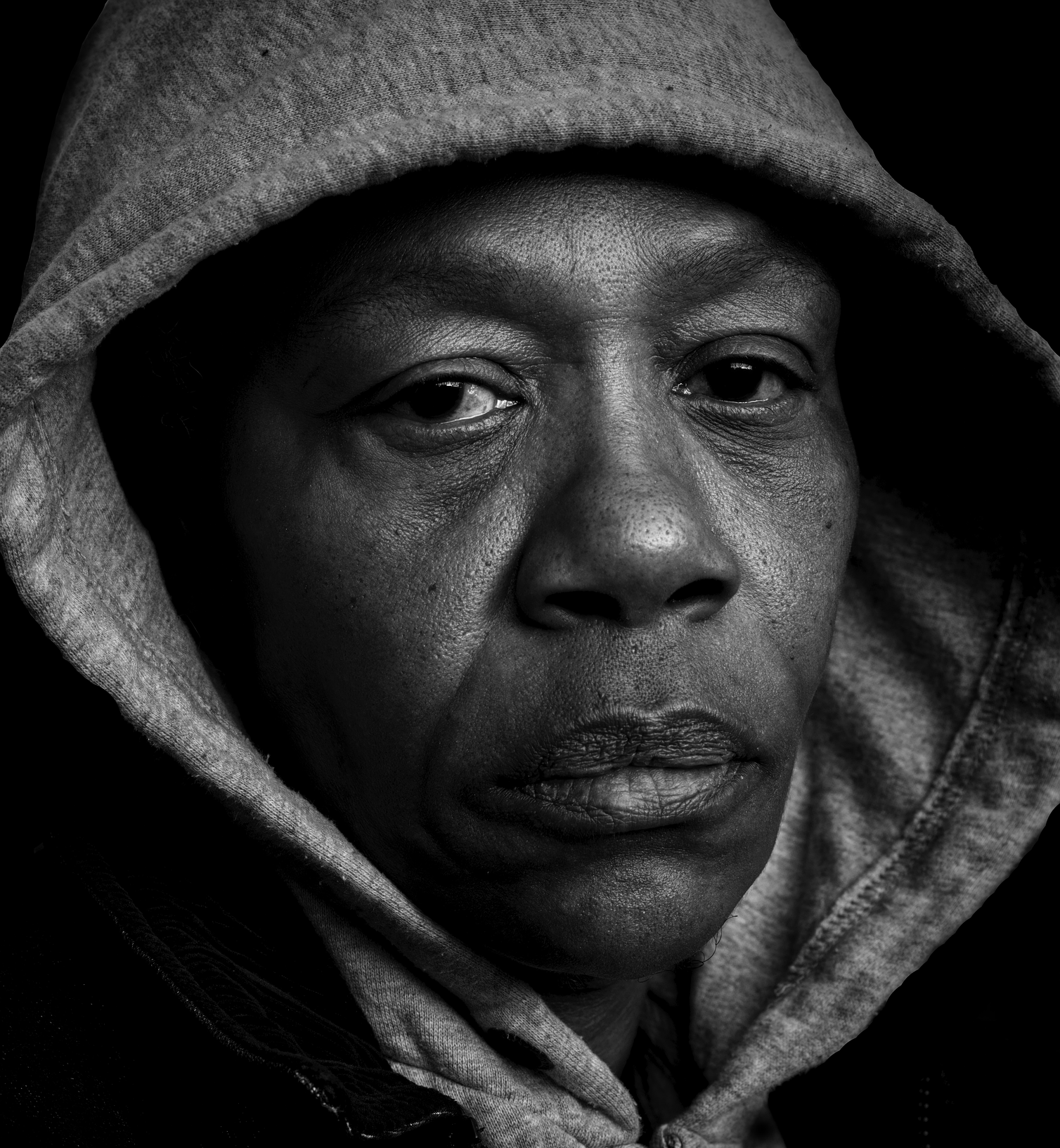 Close-up black and white photograph of a person with a hooded sweatshirt, showing their face with detailed skin texture and a serious expression.