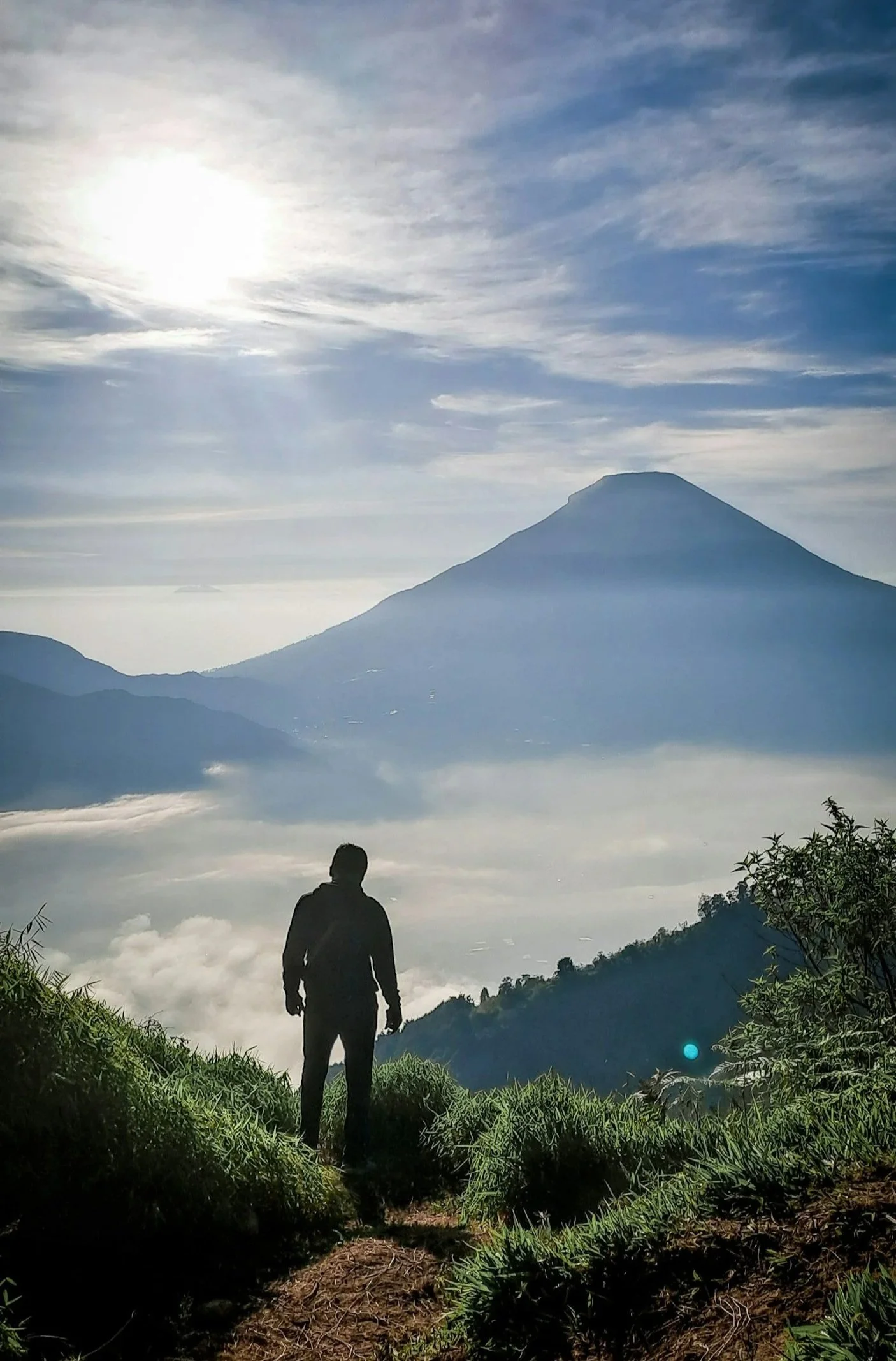 A person standing on a mountain trail with lush green grass and bushes, overlooking a valley filled with clouds. In the background, a large mountain and a prominent volcanic peak are visible under a partly cloudy sky with the sun shining brightly.