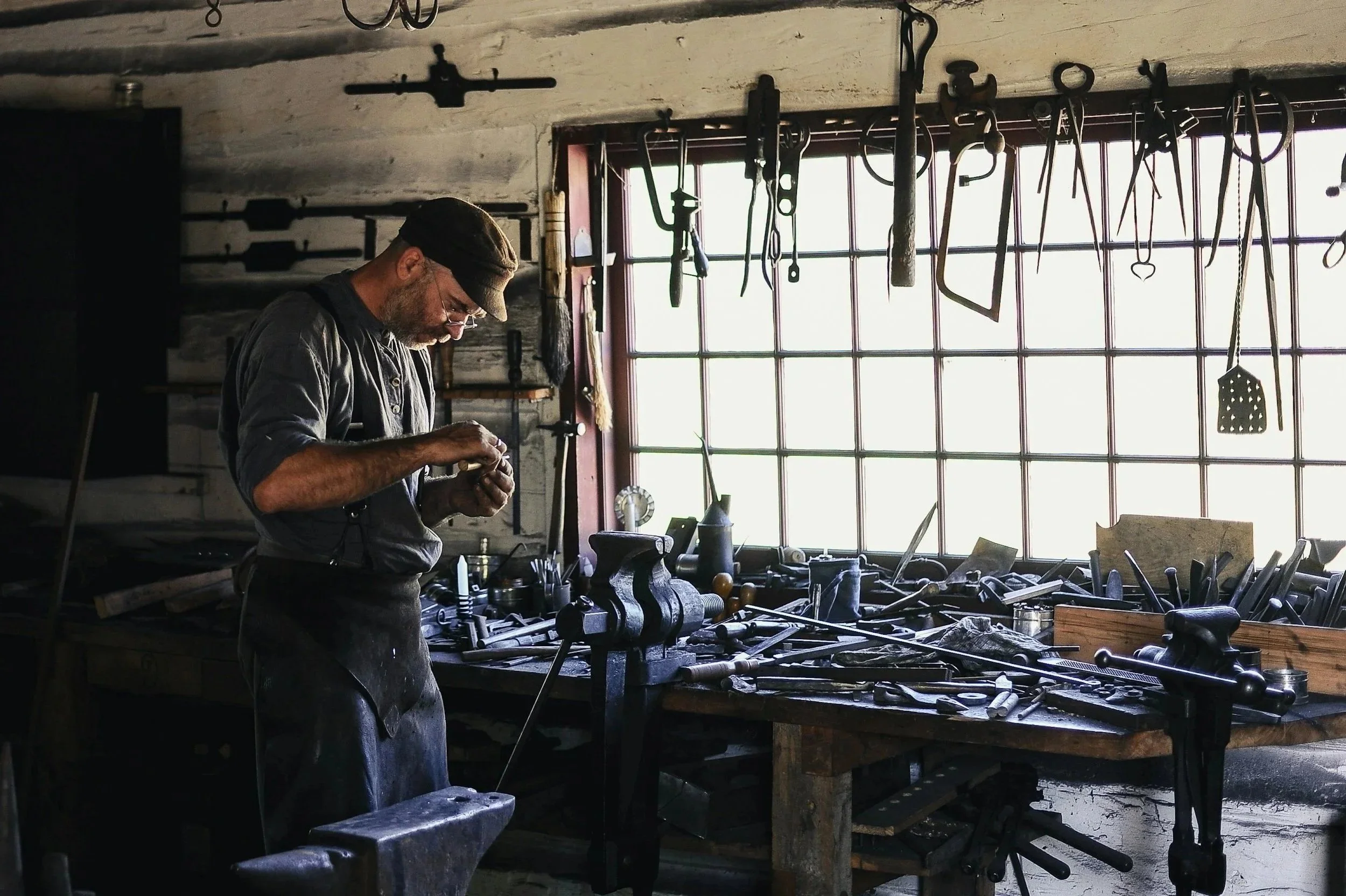 A man working in a metal workshop with various tools hanging from the ceiling, working at a workbench near a large window.