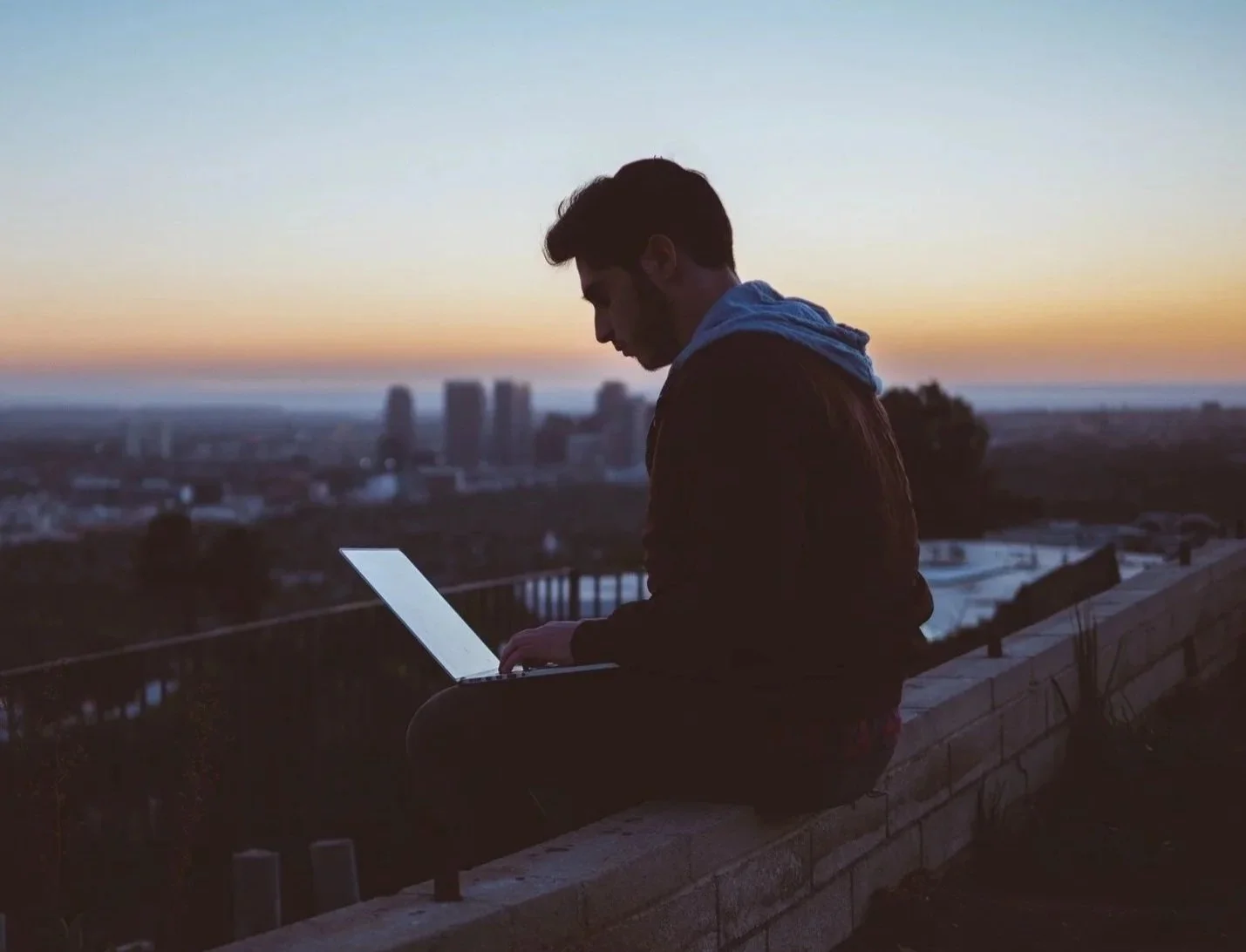 A young man sitting on a brick wall at sunset, working on a laptop with a cityscape in the background.