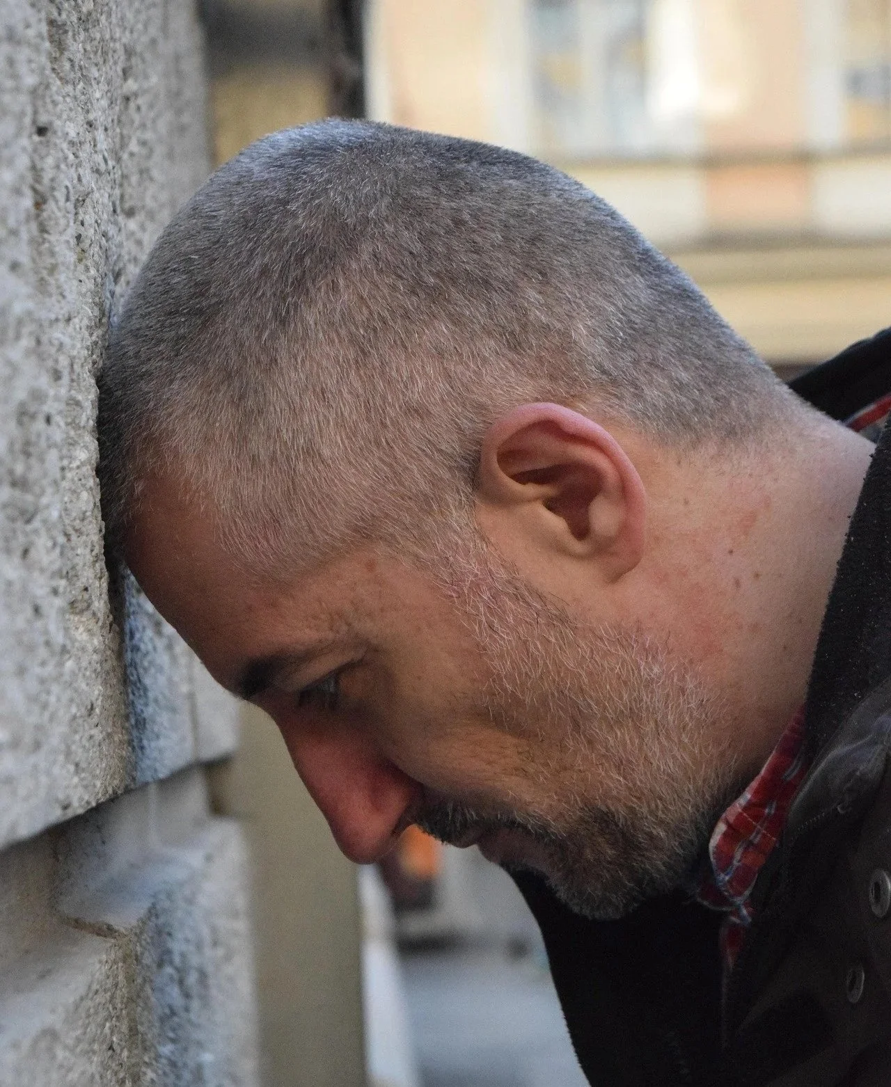 A man with grey hair and beard presses his forehead against a brick wall with his eyes closed.
