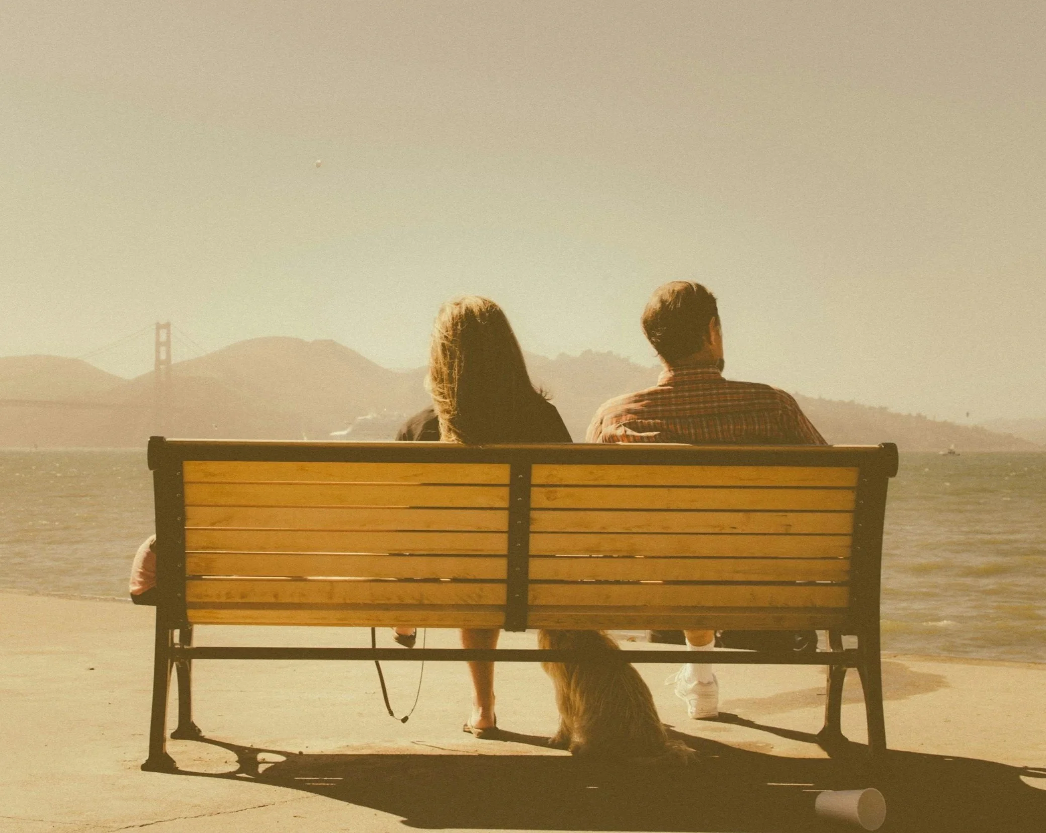 A couple sitting on a park bench facing the water with the Golden Gate Bridge in the background, a dog standing beside the bench, and a person white sneakers visible underneath the bench.