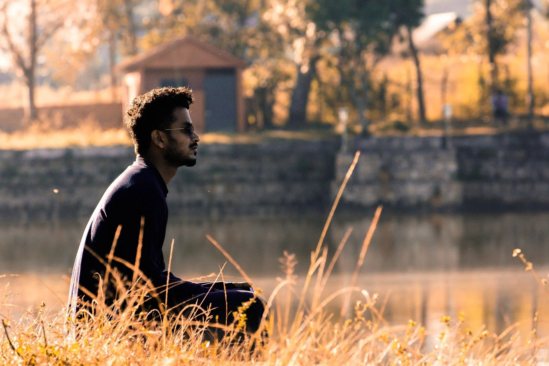 A man with sunglasses and a beard sitting outdoors among tall grass and plants, with trees and a small wooden building in the background during sunset or late afternoon.