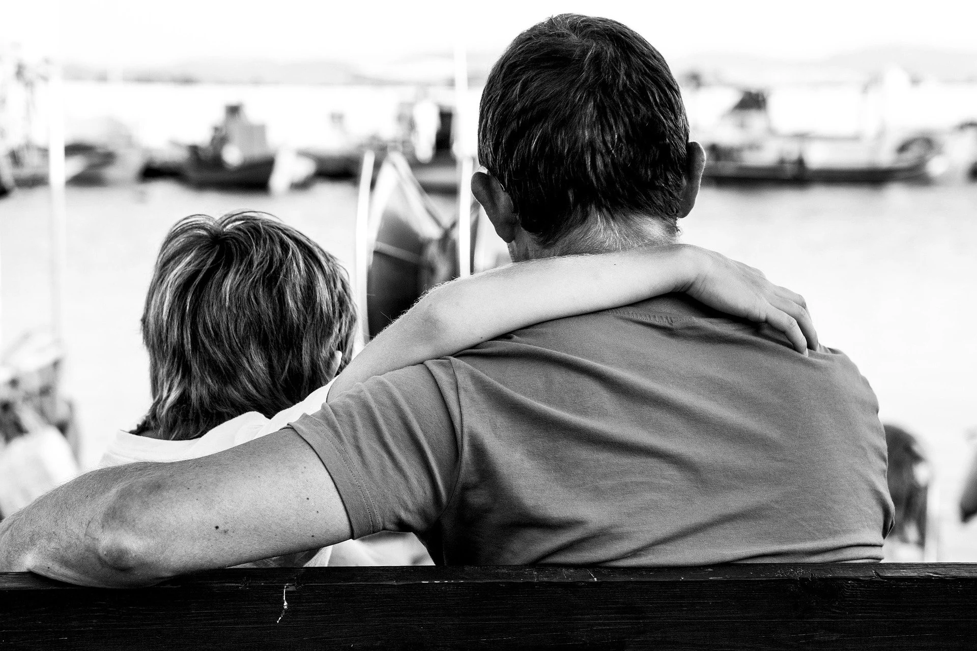 A man and a woman sitting closely together with their arms around each other, looking towards boats docked on the water, in a black and white photo.