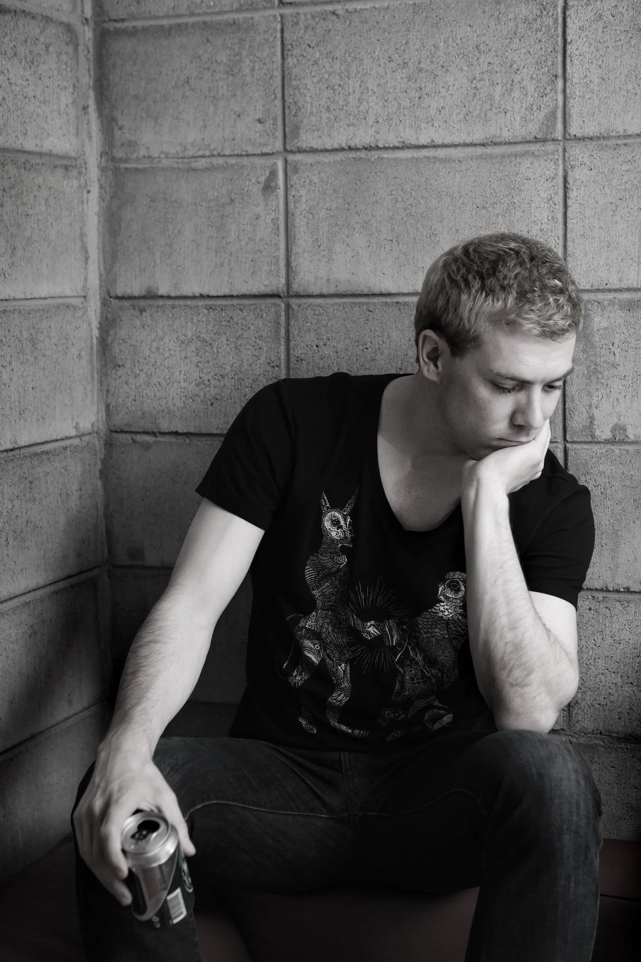 A young man with short curly hair and a black T-shirt featuring animal illustrations sits against a brick wall, looking down with a thoughtful expression, holding a can in his right hand.
