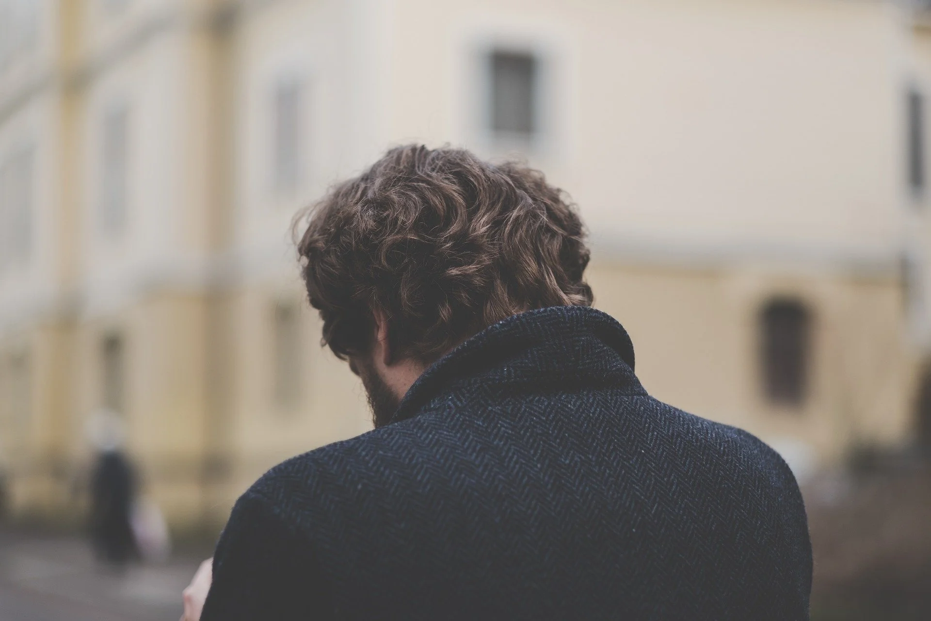 Back view of a man with curly hair wearing a dark coat, standing outdoors.