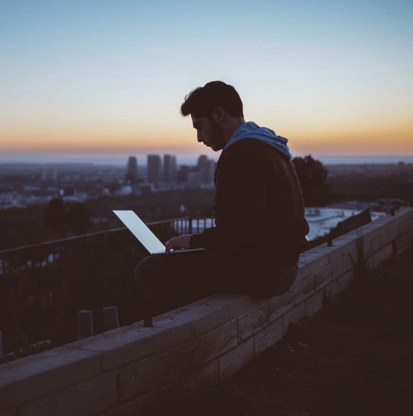 A young man sitting on a brick wall outdoors at sunset, using a laptop with a cityscape and a colorful sky in the background.