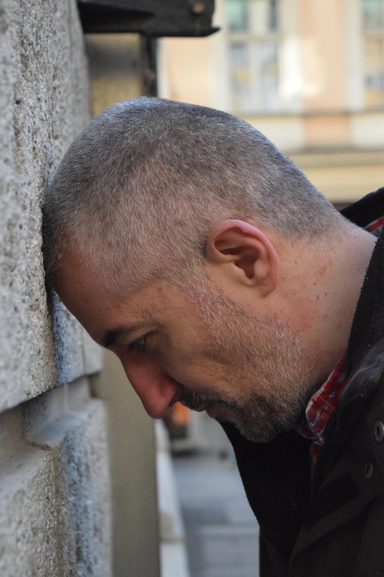 A man with a short gray haircut and beard resting his forehead against a textured brick wall, eyes closed, outdoors in an urban environment.