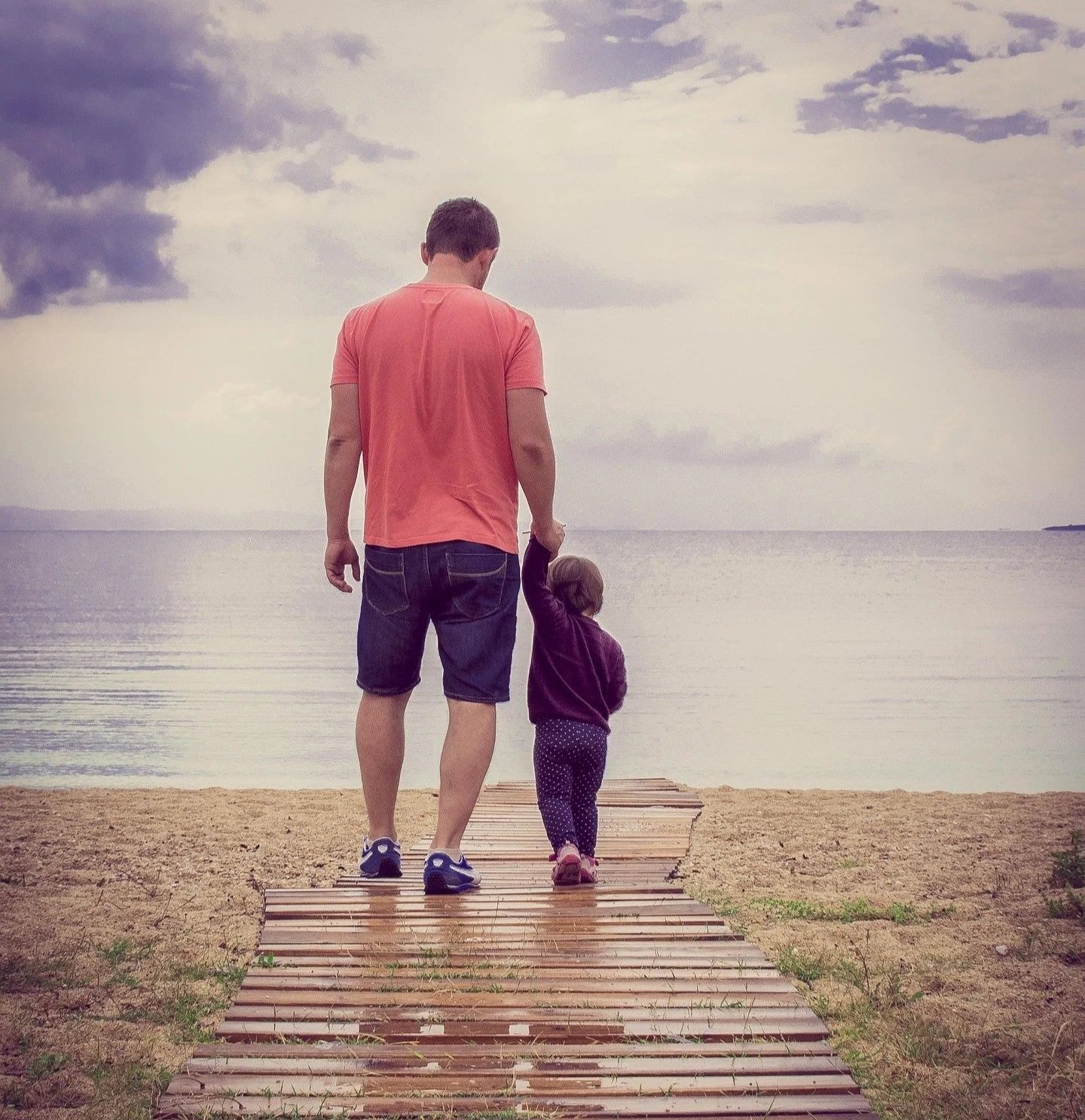 A man and a young girl walking on a wooden path towards the water, holding hands at the beach during cloudy weather.
