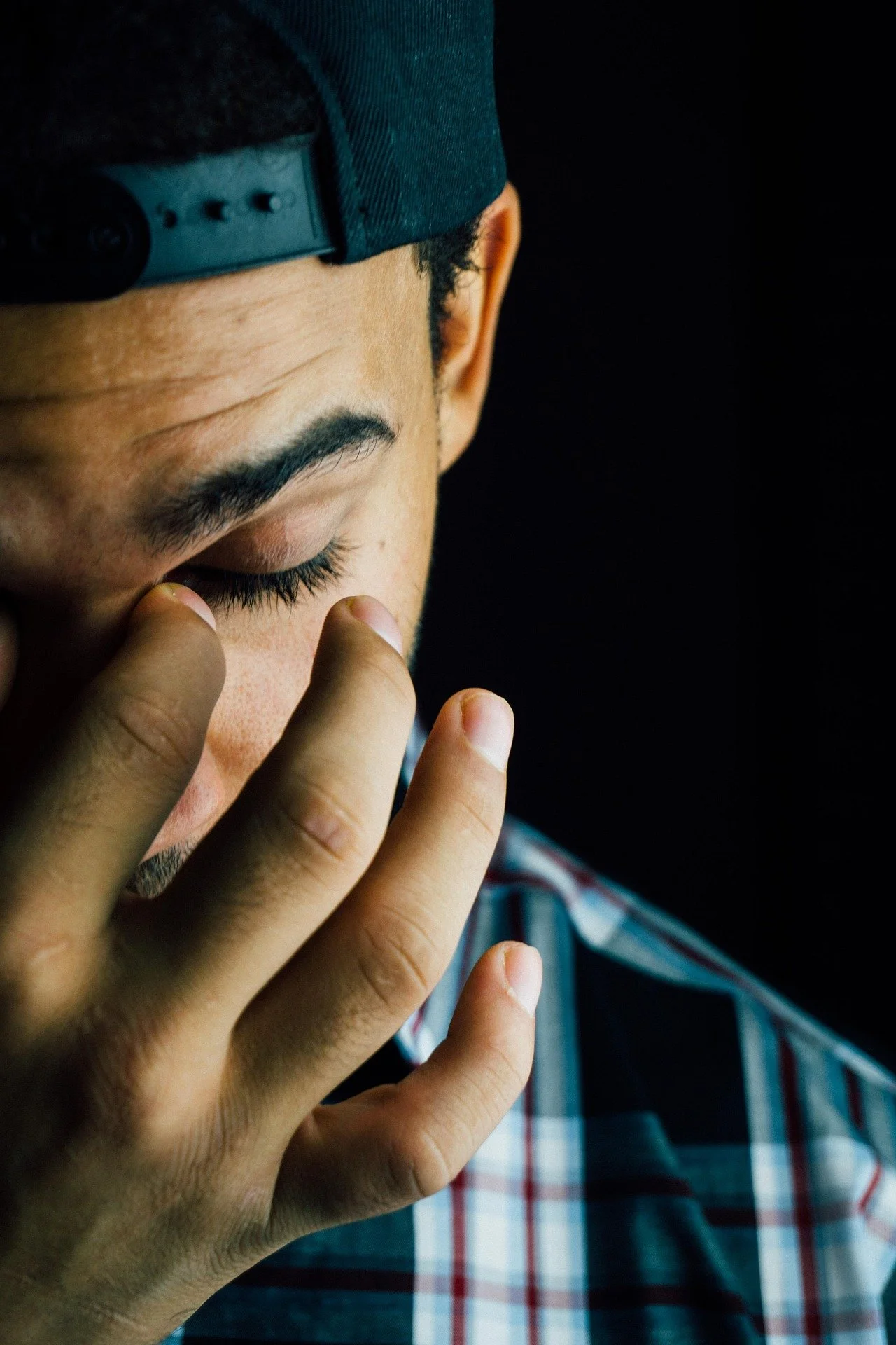 A person with a black cap and plaid shirt, holding their hand near their face with eyes closed.