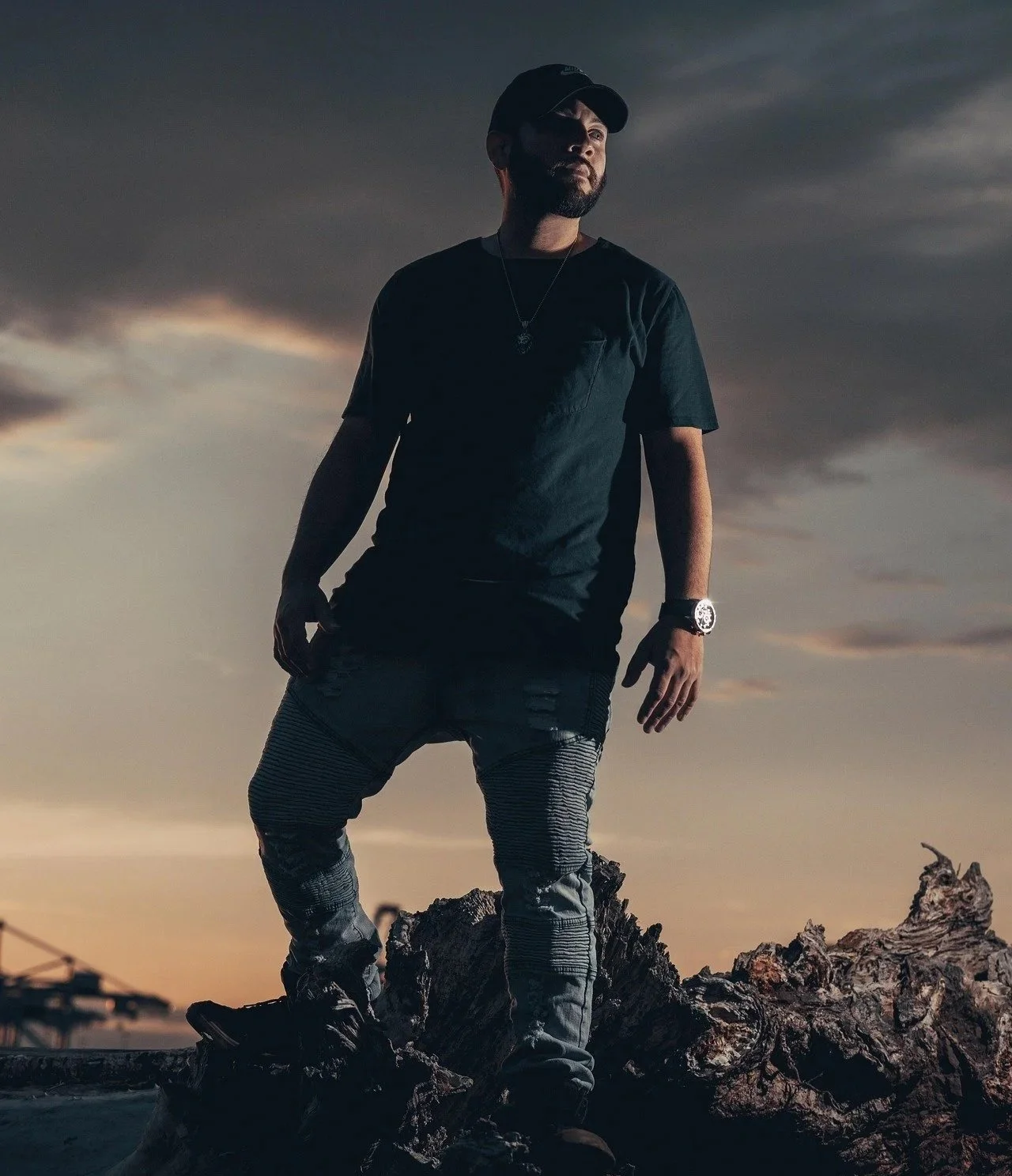 A man standing outdoors on a large piece of driftwood at sunset, wearing a black T-shirt, striped pants, a baseball cap, and watch, with a dark sky background.