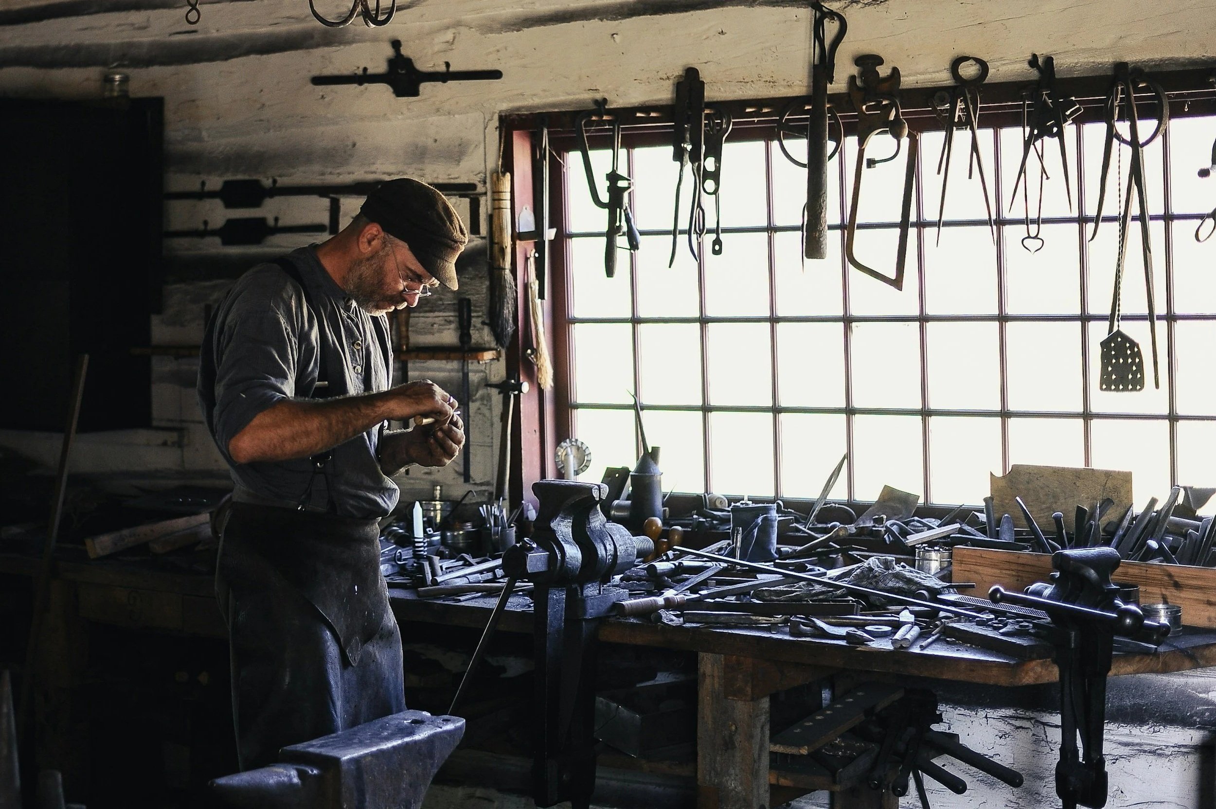 A man working in a vintage woodworking shop, surrounded by tools hanging from the ceiling and placed on the workbench, with natural light coming through a large window.