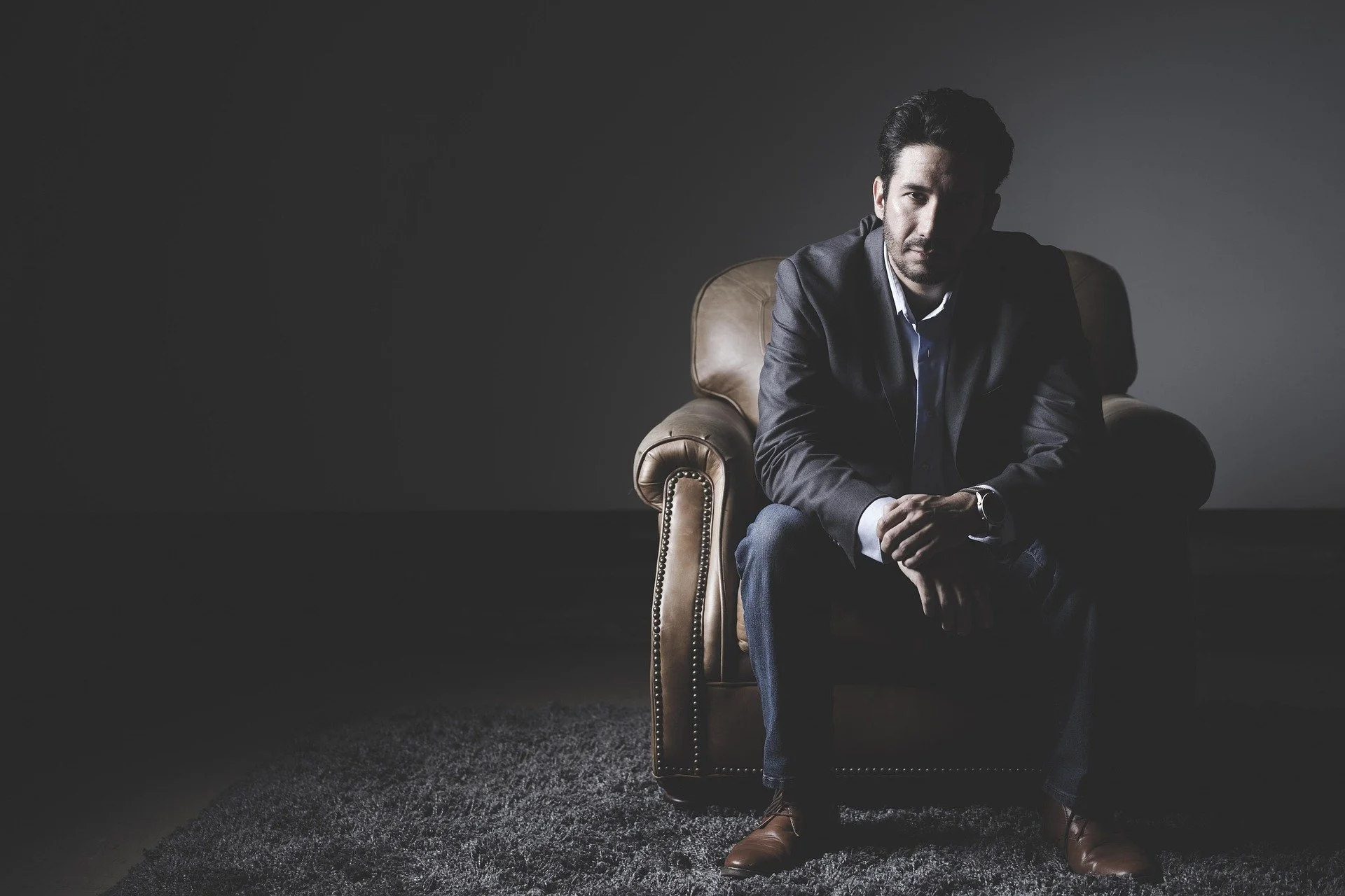 A man in a dark suit sitting on a brown leather armchair in a dimly lit room with a dark gray background.