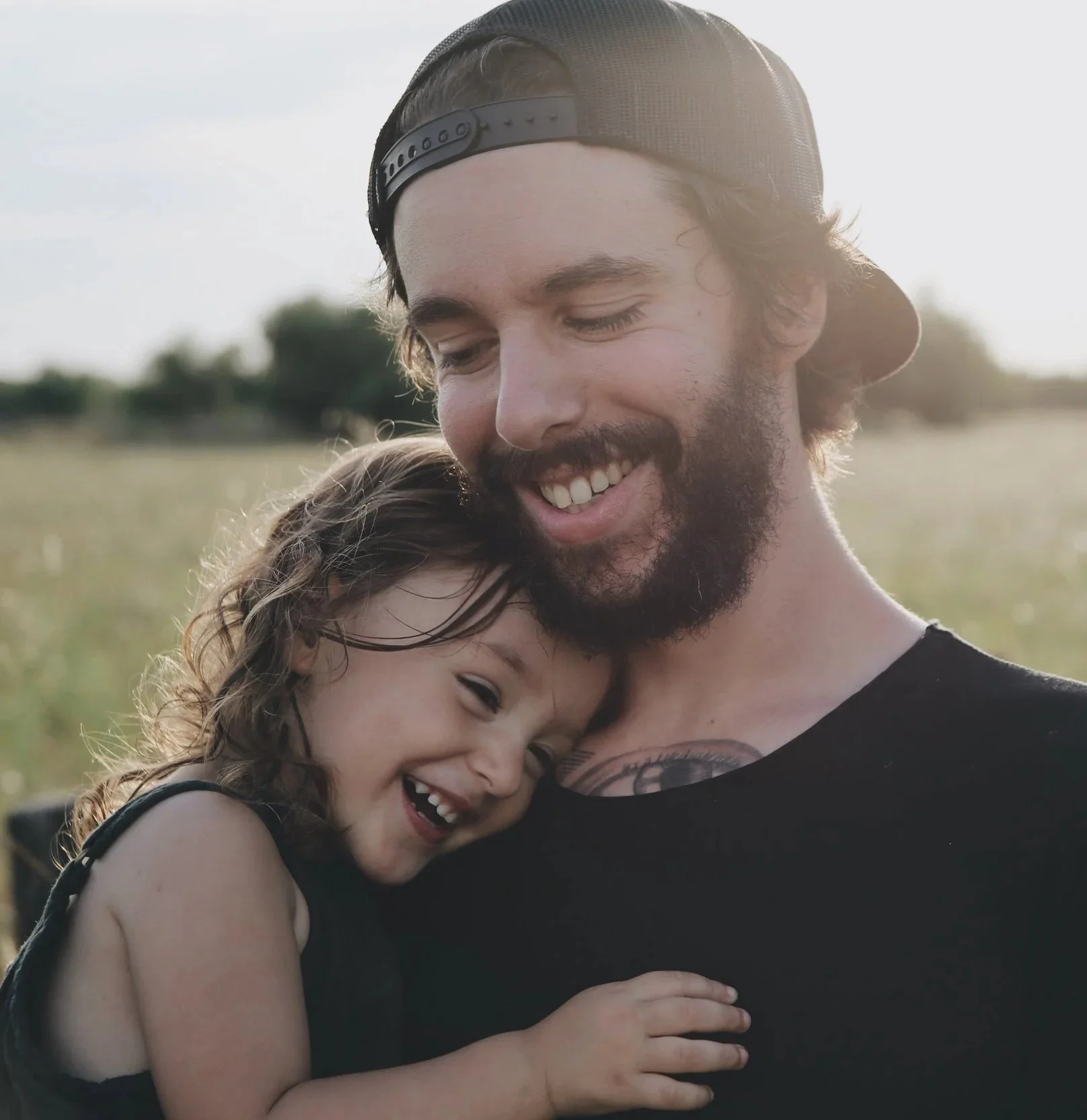 A man with a beard, wearing a black cap backwards, smiling and hugging a young girl with curly hair, outdoors in a field.