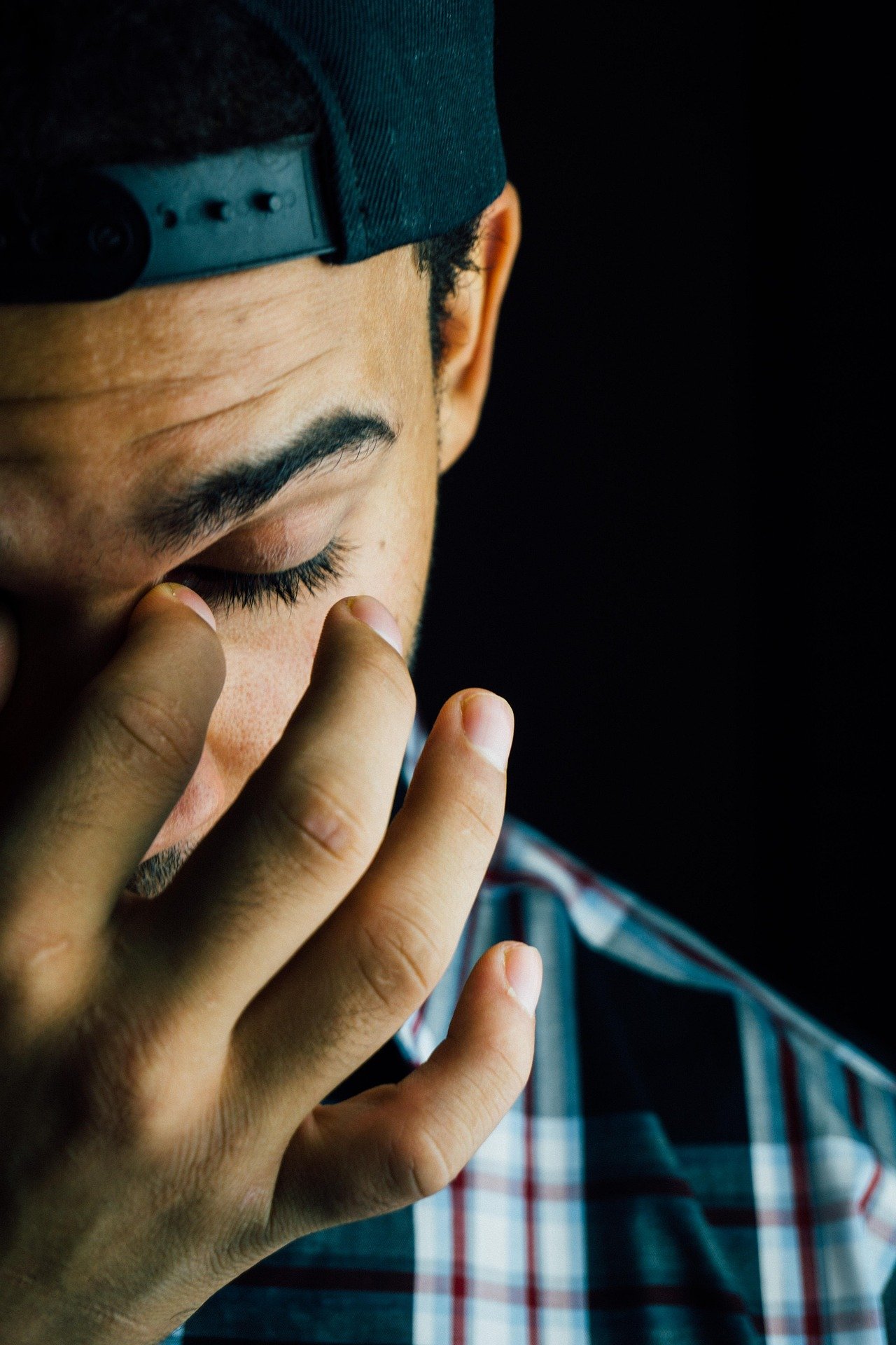 A person with curly hair wearing a blue quilted cap and a plaid shirt, touching the side of their face with fingers, against a black background.