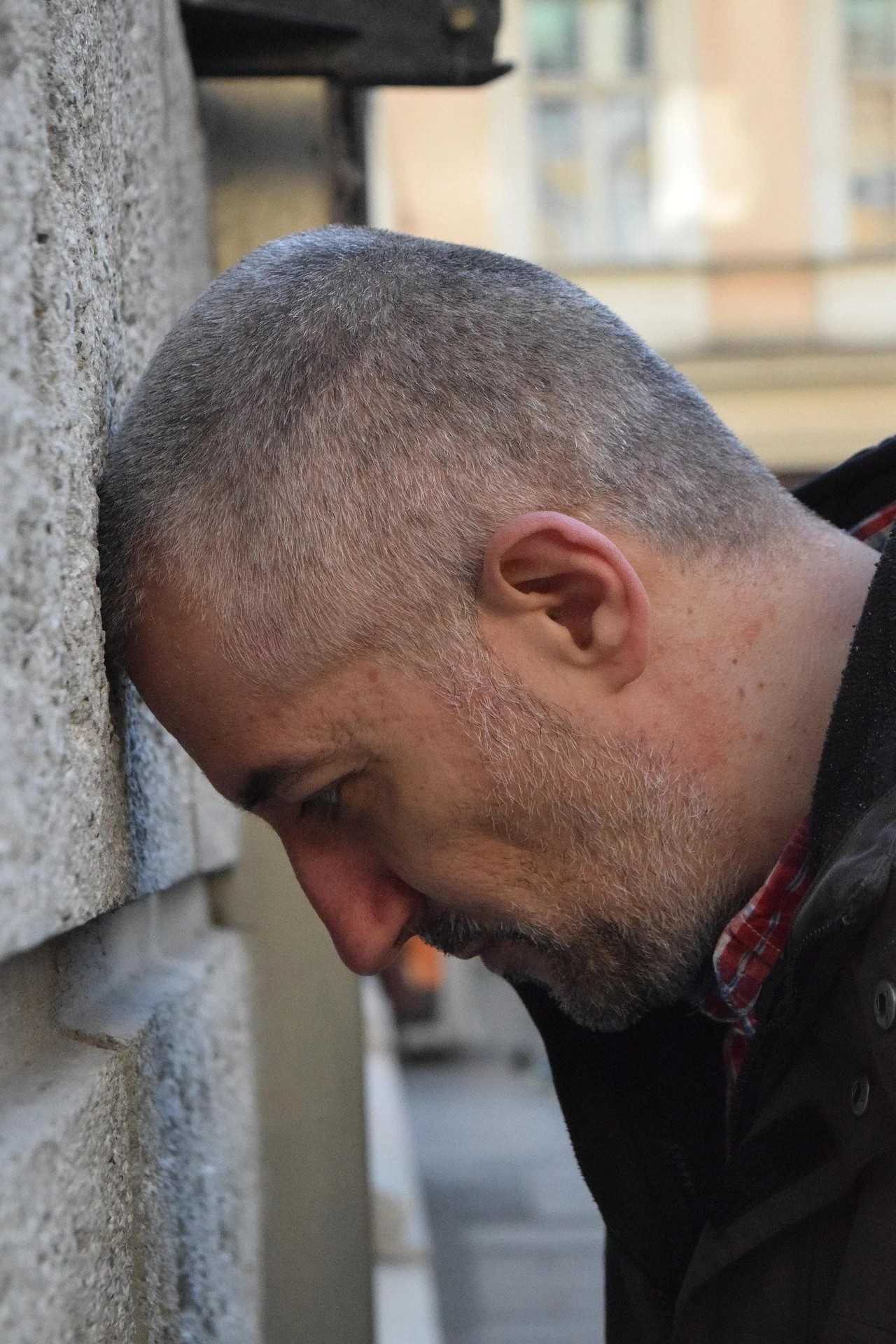 A man with a closely shaved head and beard is leaning his head against a stone wall with his eyes closed.