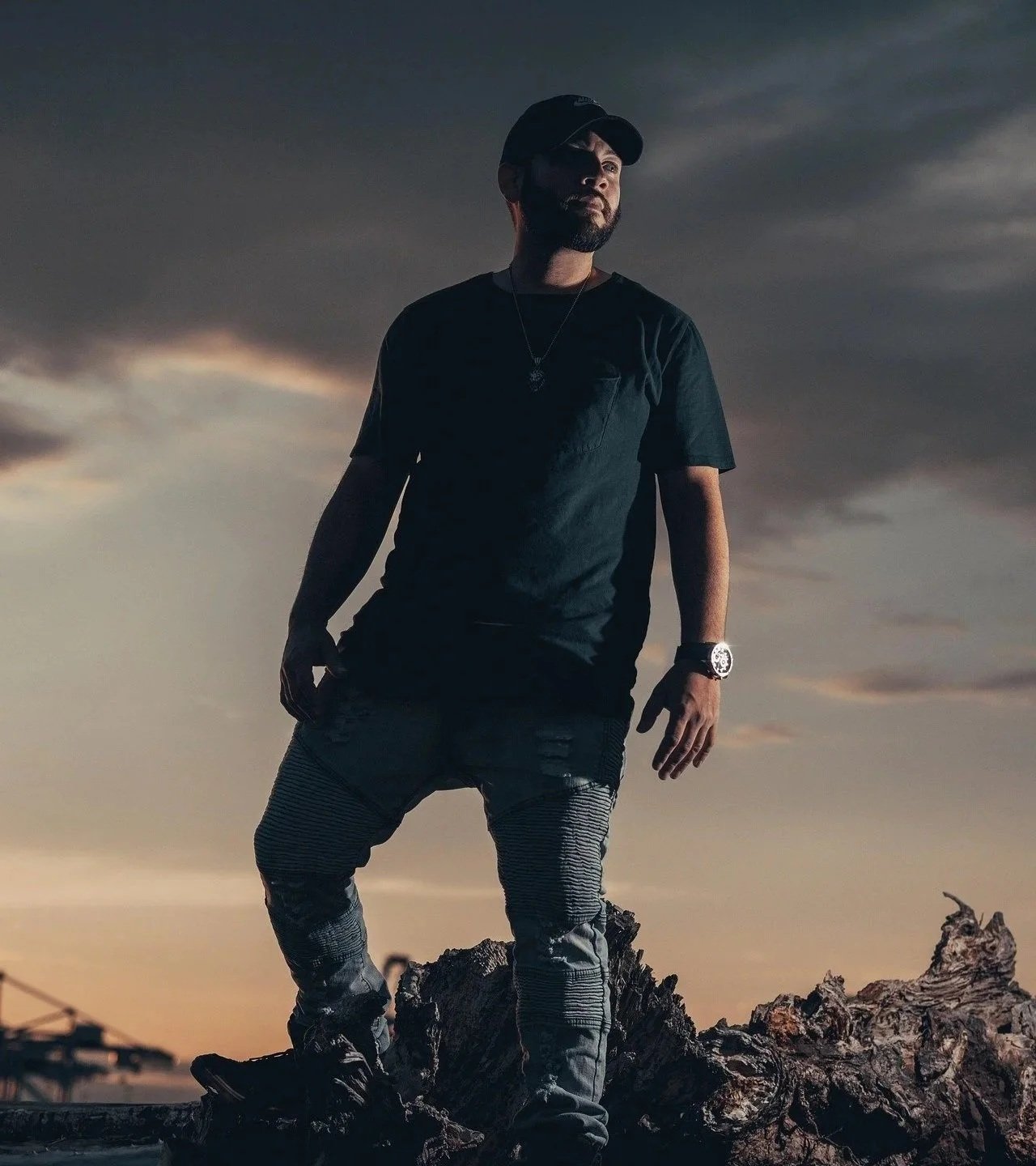 A man standing outdoors on a piece of driftwood during sunset, wearing a black T-shirt, striped pants, a cap, a watch, and a necklace, with cloudy sky background.