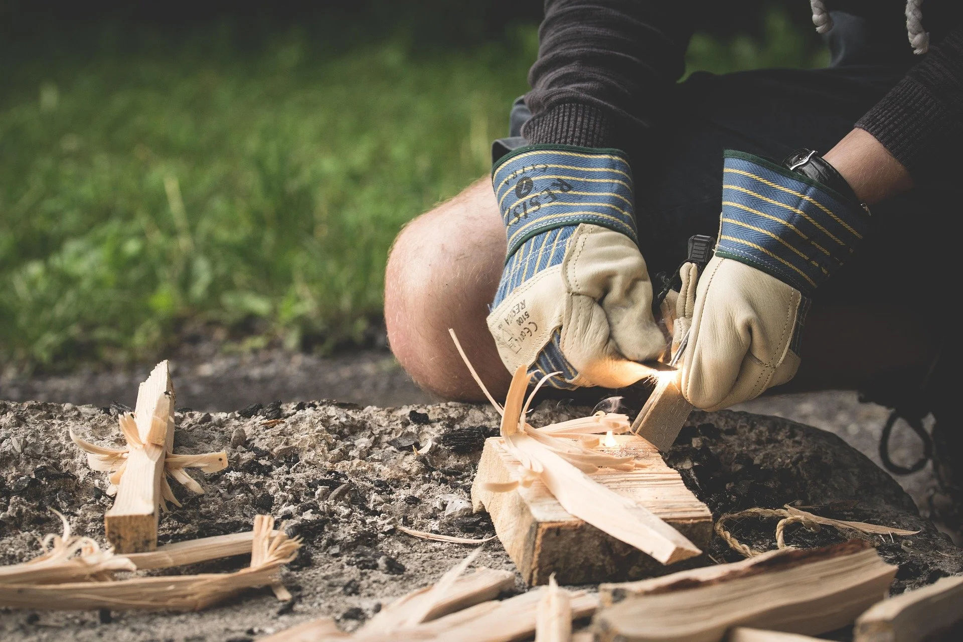 A person wearing protective gloves and a race wristband using a chisel and hammer to carve wood on a tree stump outdoors, with wood shavings and wood pieces on the ground.