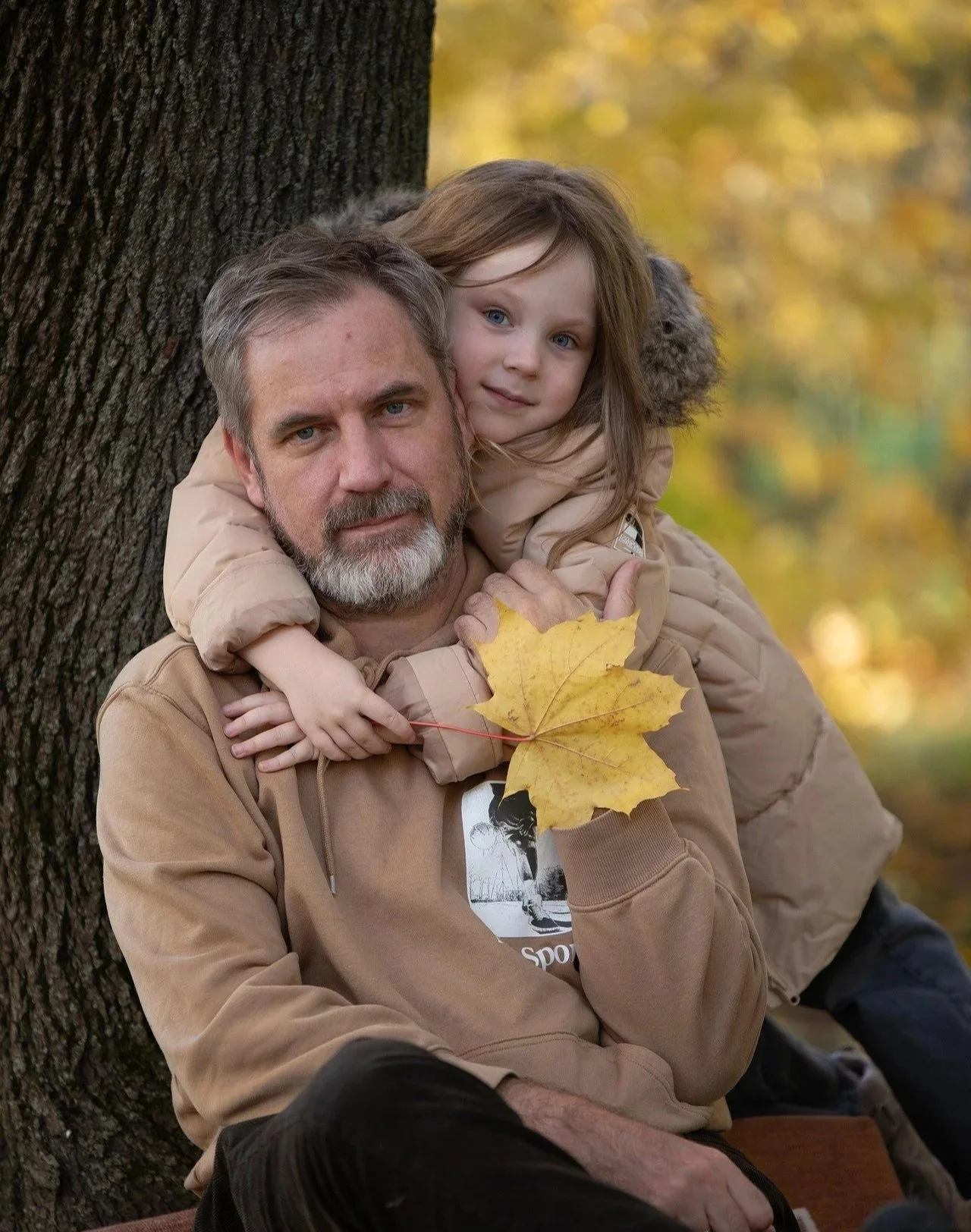 A man with gray hair and a beard holding a young girl on his shoulders, both wearing beige jackets, outdoors in autumn. The girl has light brown hair and blue eyes, and they are near a large tree with yellow leaves. The man is holding a yellow maple leaf.