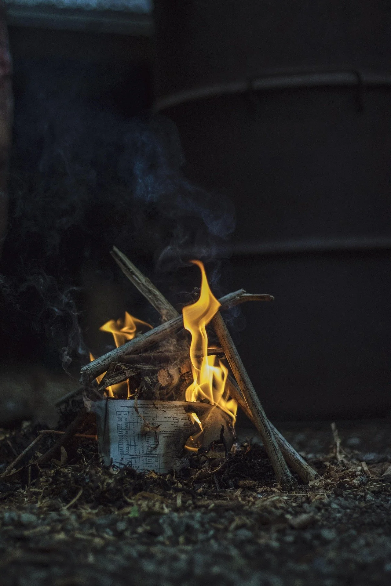 Small fire made with sticks and paper on the ground, with flames and smoke rising in a dark setting.